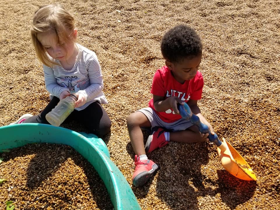 Two children playing in wood chips, one with a bottle, one with a shovel