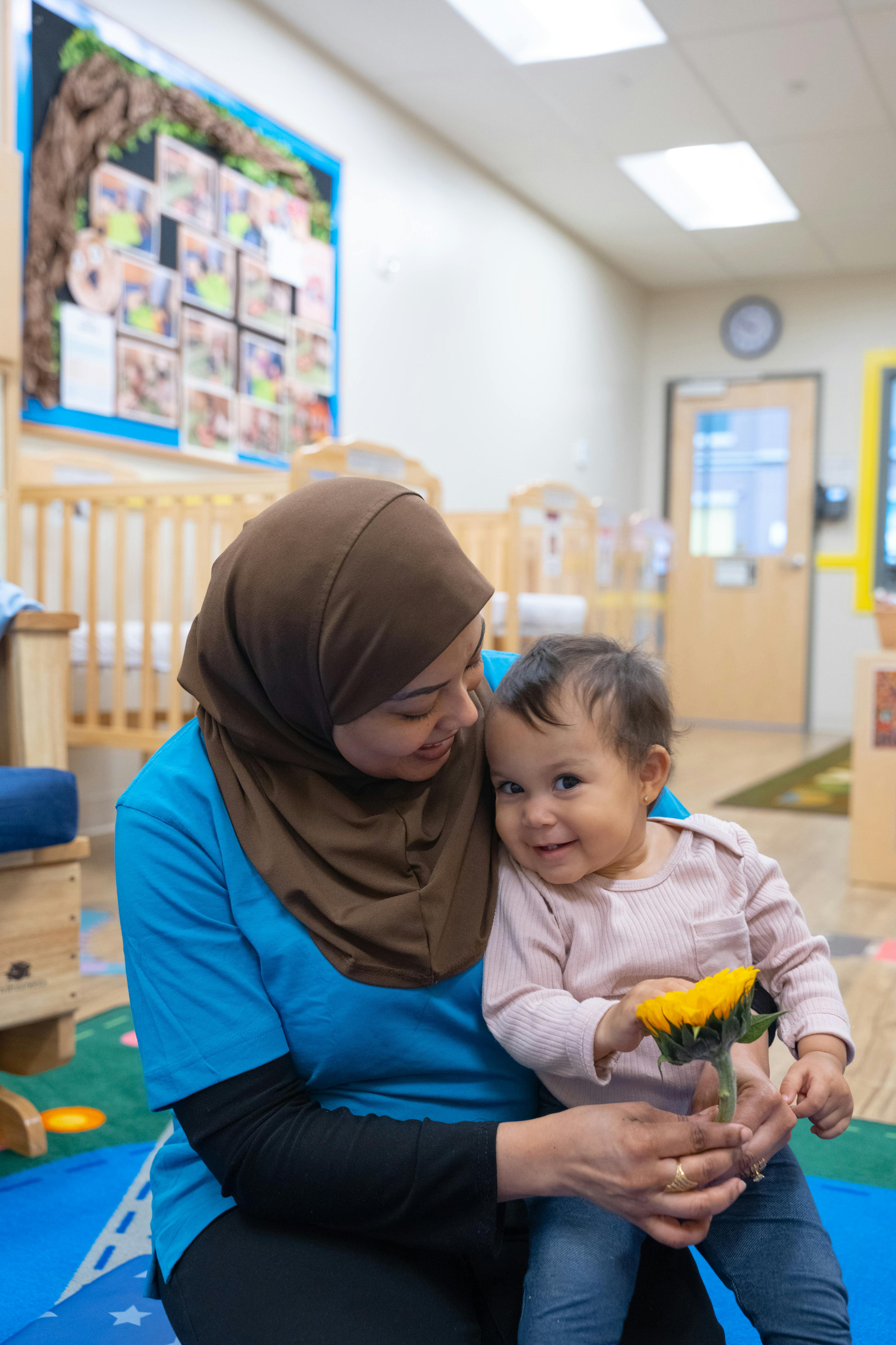 Person in hijab holding a child with a flower in a classroom
