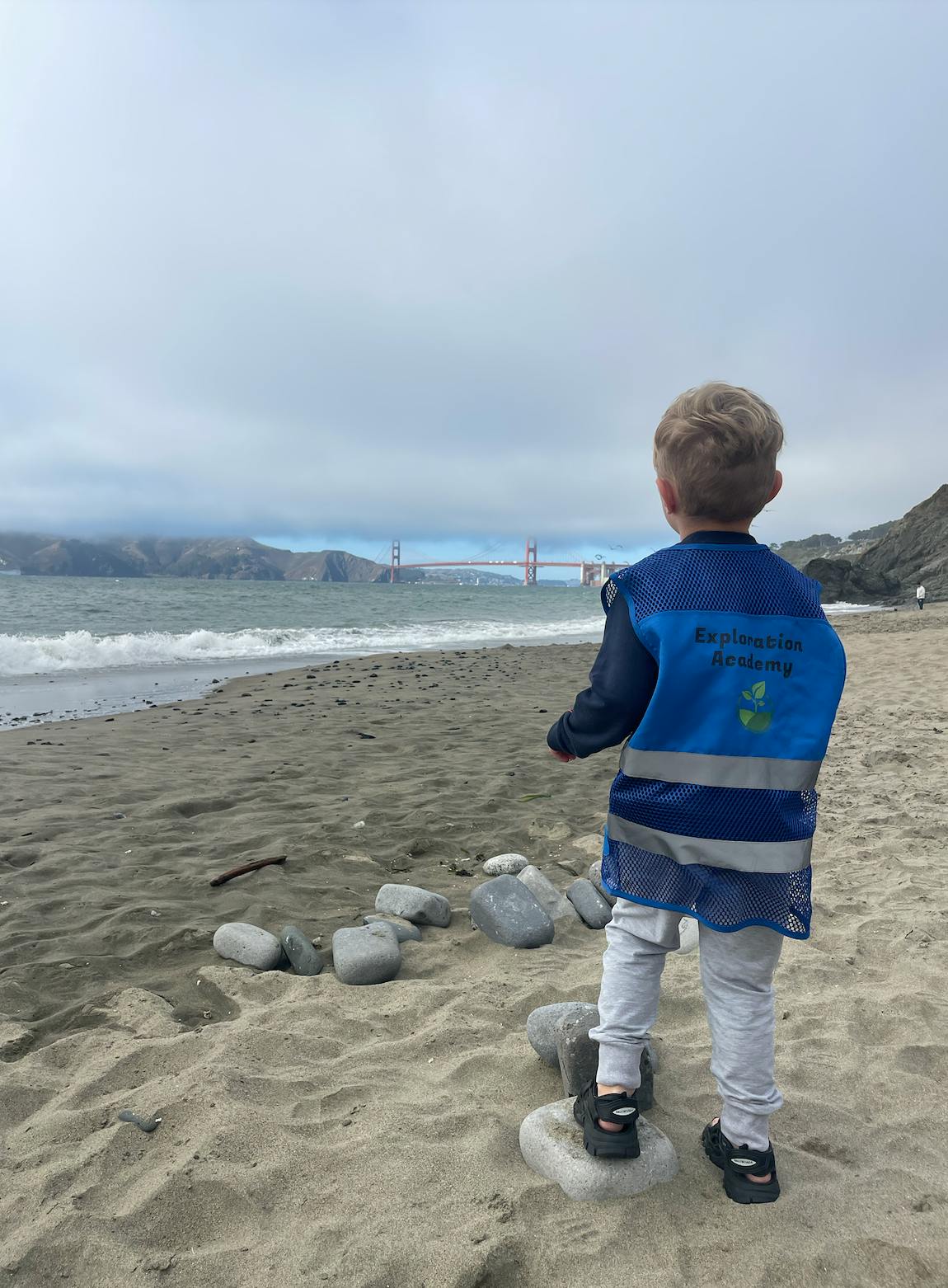 Child in a blue vest standing on a rock on a sandy beach looking at the ocean