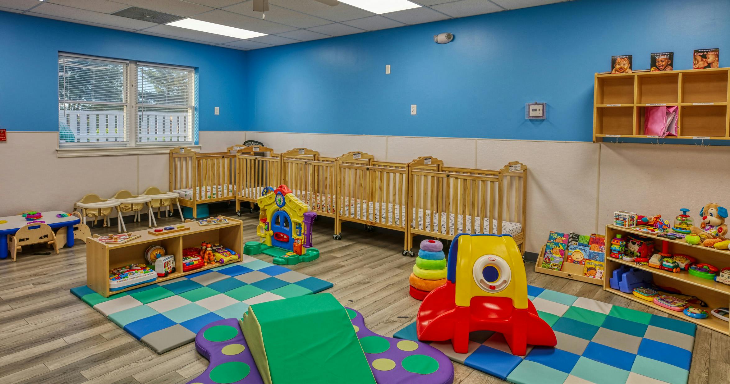 A colorful daycare room with several wooden cribs and various infant toys