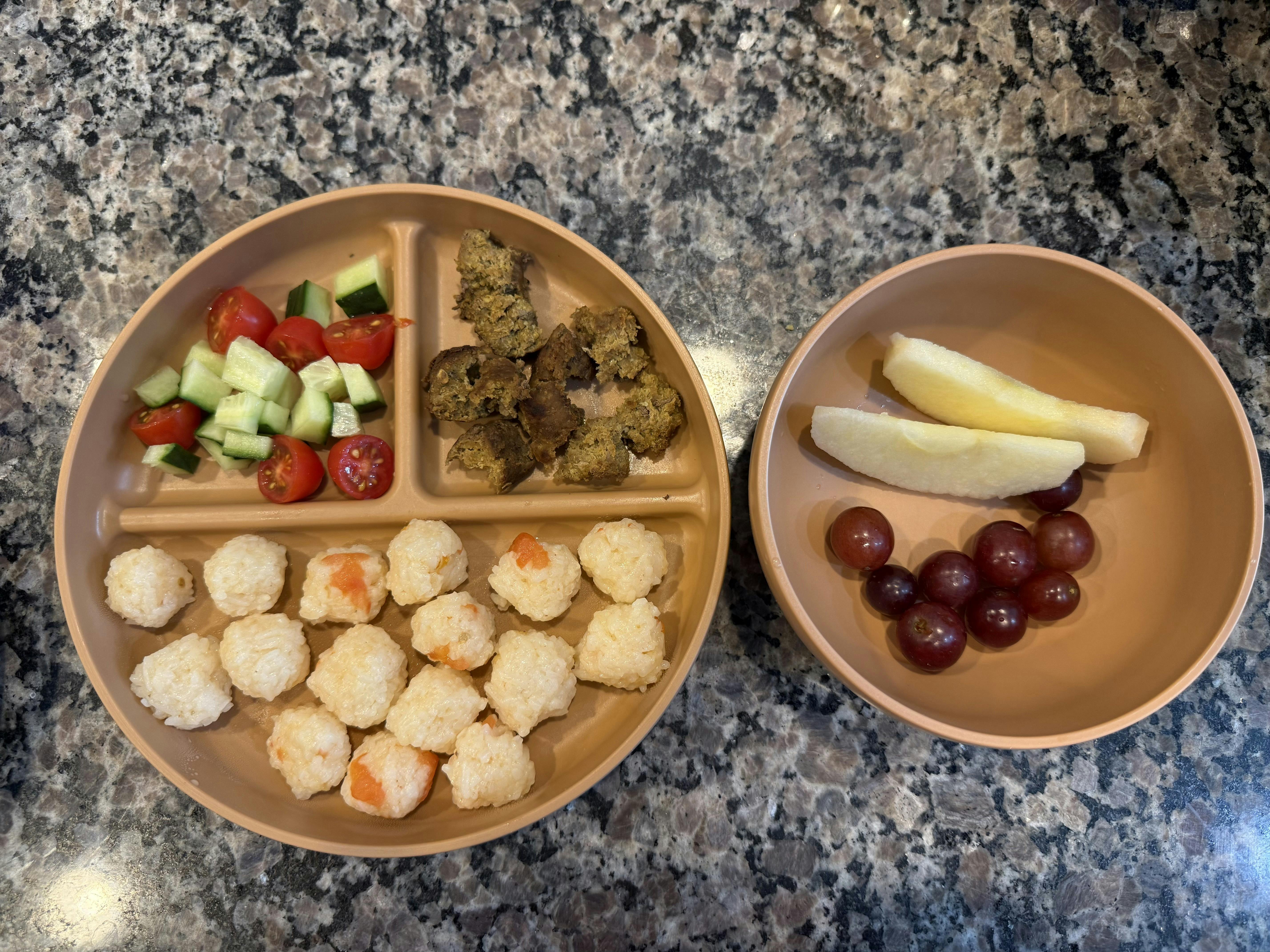 A beige divided plate with cucumber, tomato, meatballs, and rice balls, plus a bowl of apple slices and grapes