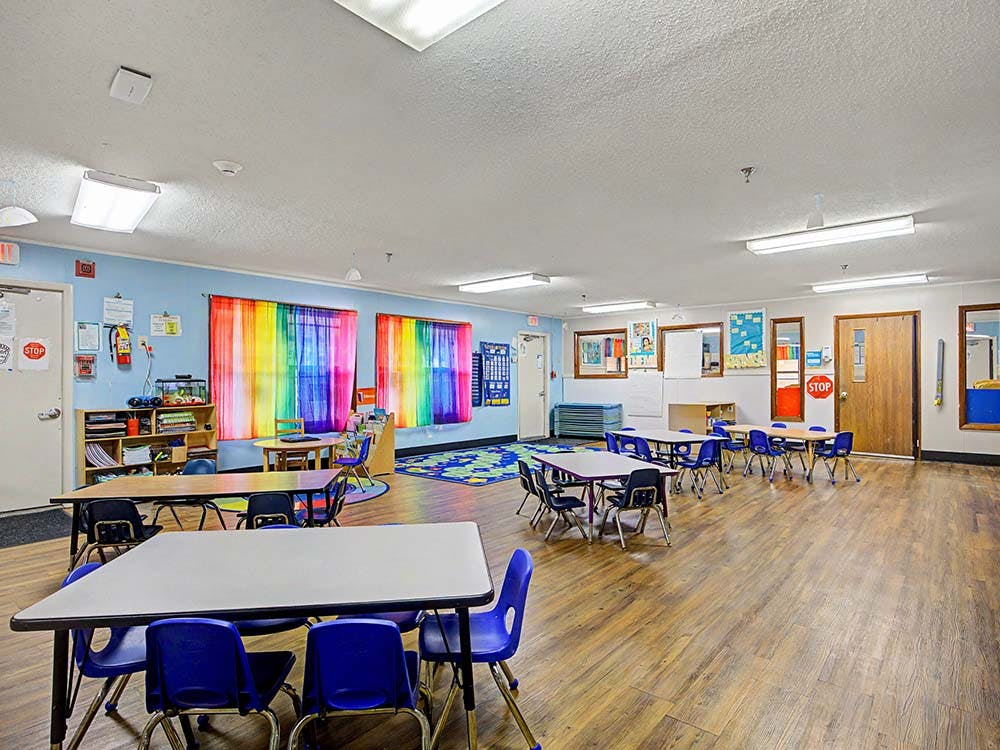 Classroom with small tables and chairs, colorful rainbow curtains, and a wooden floor