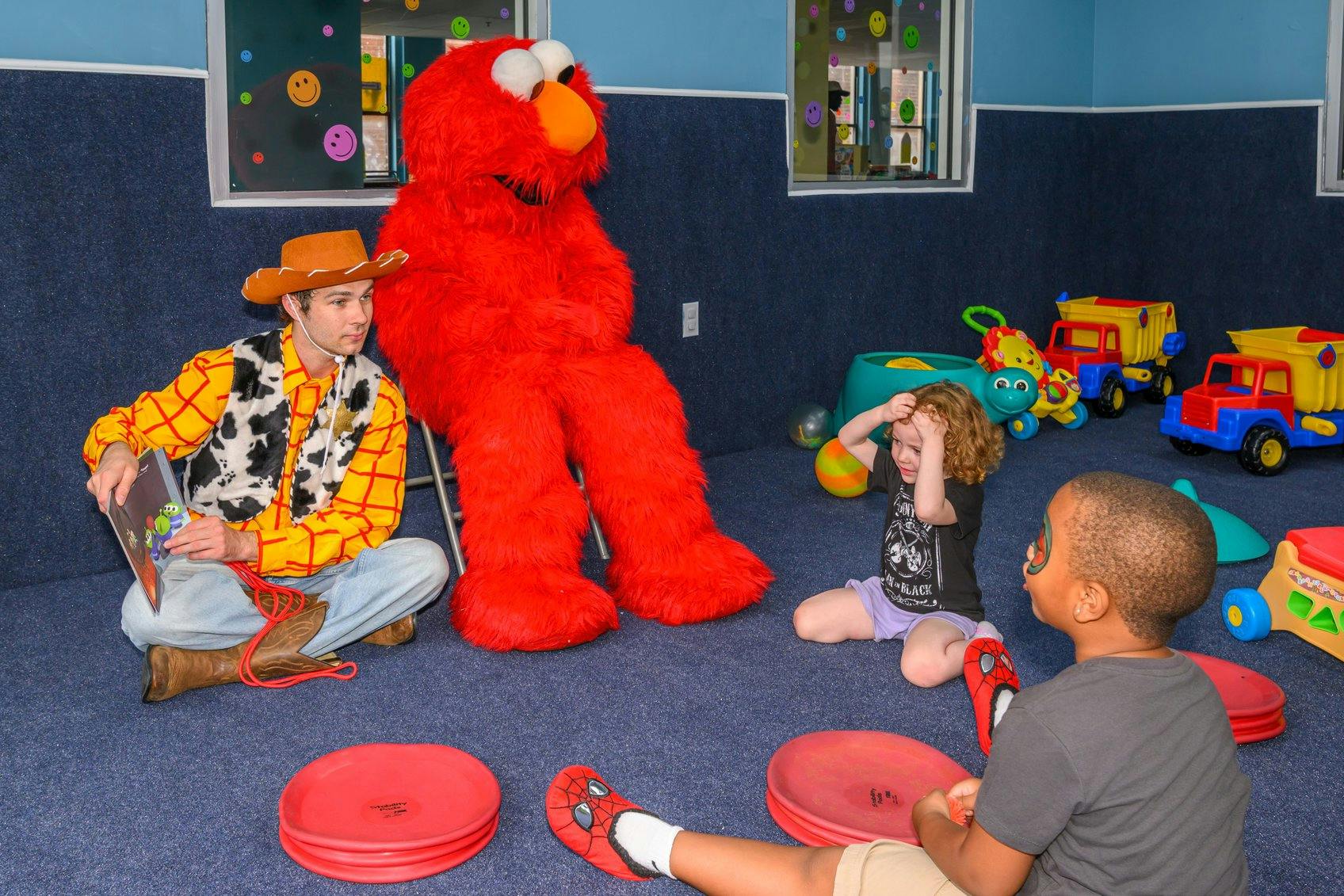 A person dressed as a cowboy reading a book to two children near a mascot