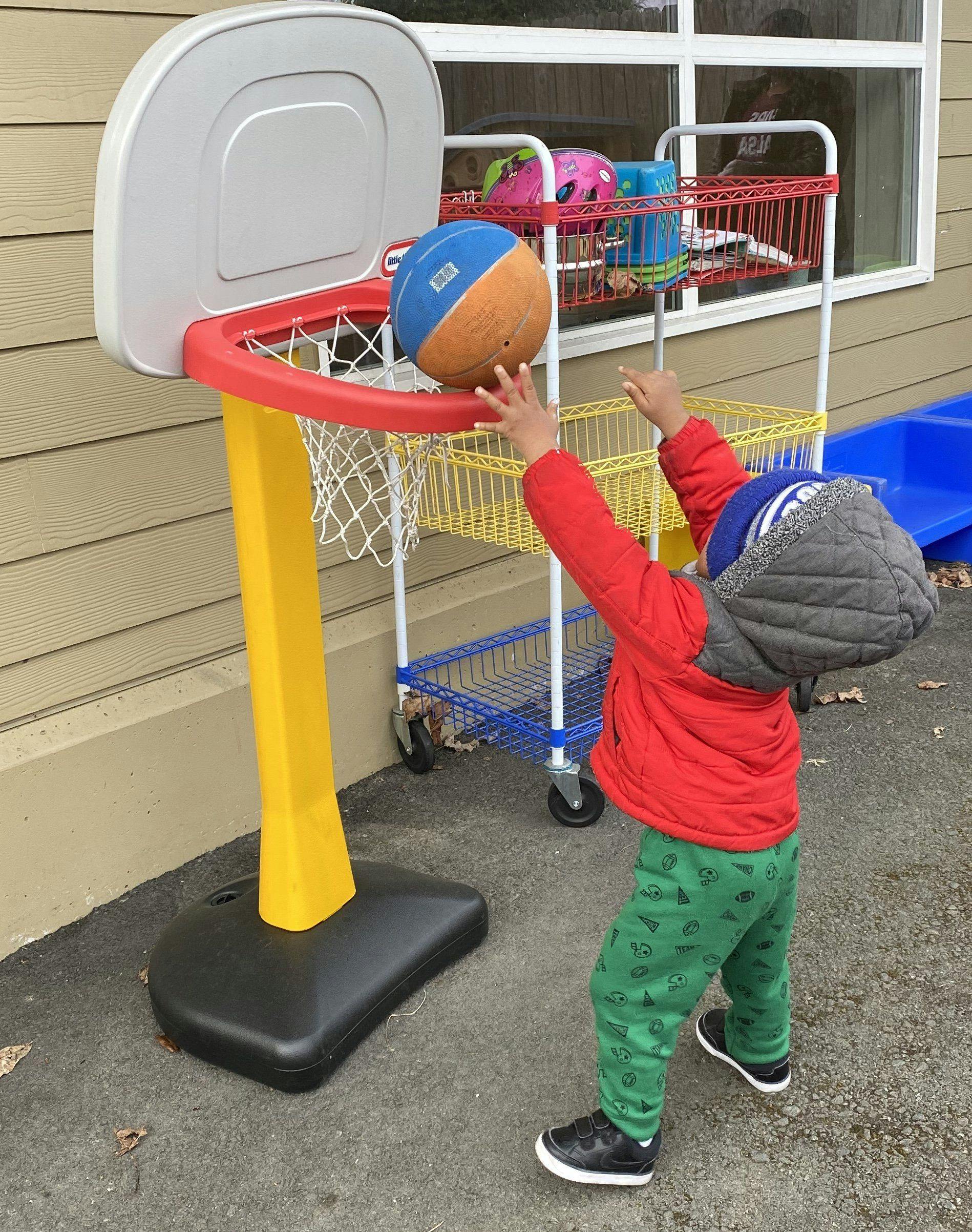 Child wearing a red jacket and green pants attempting to dunk a basketball
