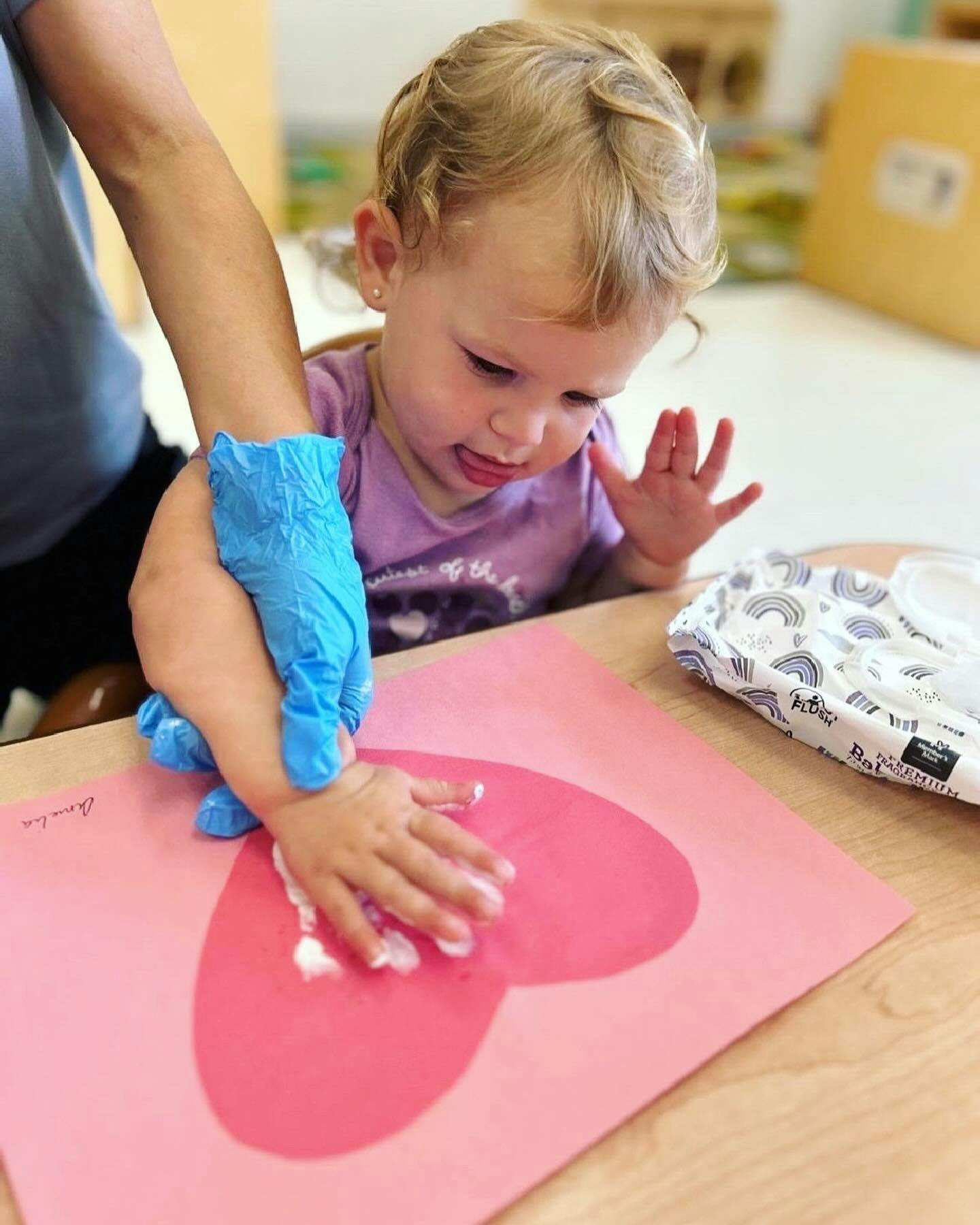 Person in blue gloves helping a child make a handprint on a pink heart