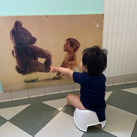 Child sitting on a potty, pointing at a poster with a teddy bear and baby