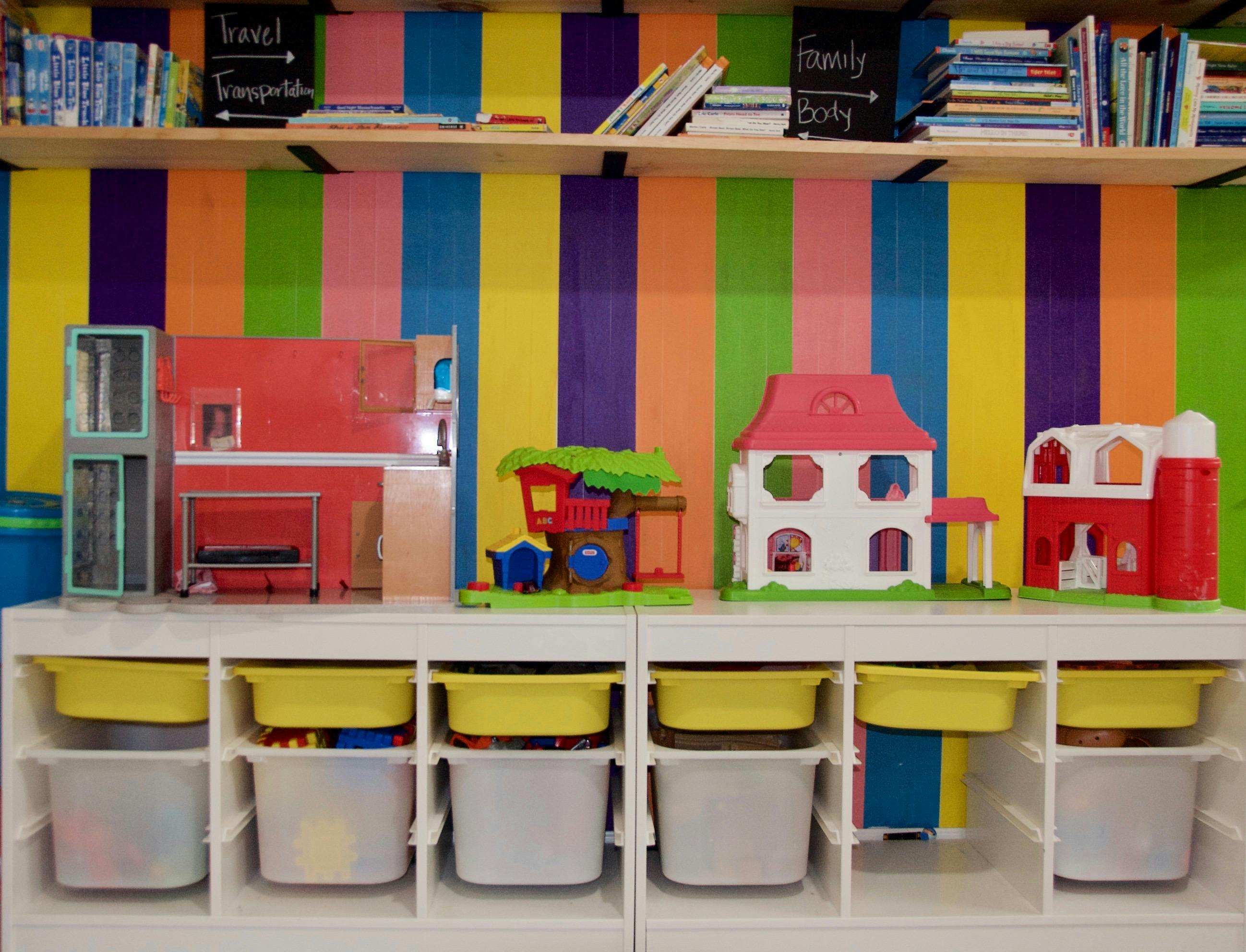 Dollhouses and toys on white shelves with books above, against a striped wall