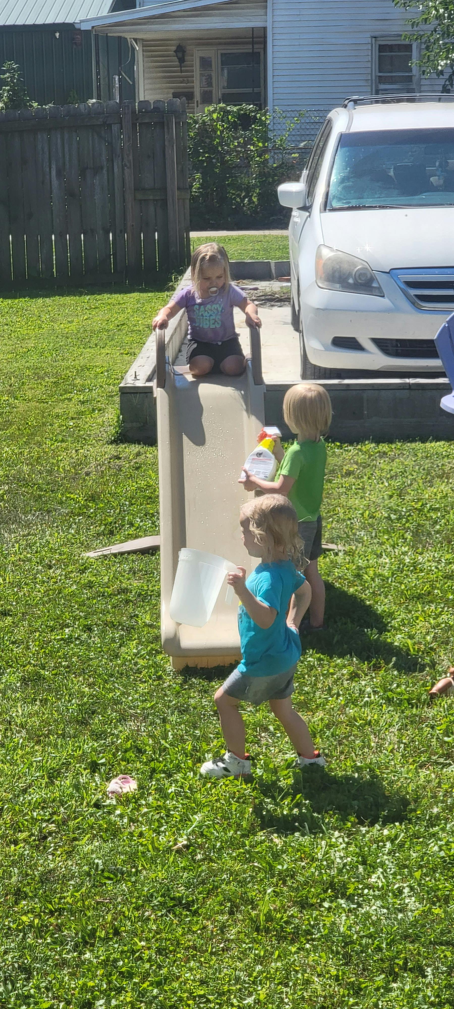 Three children playing with a slide and water containers outdoors