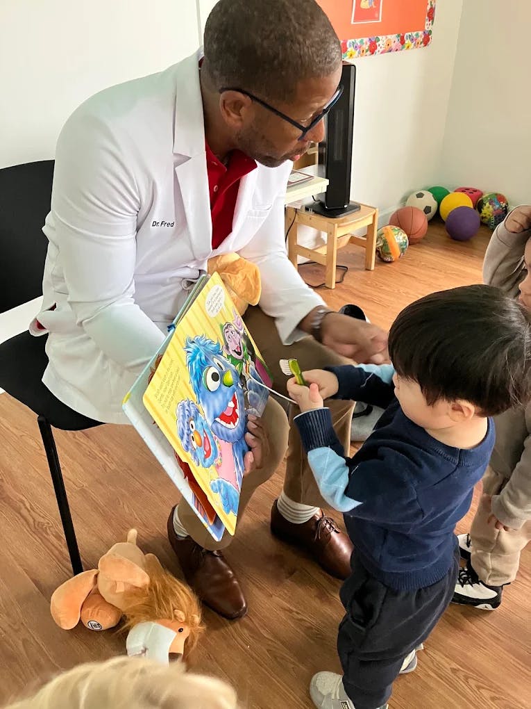 Person in white coat reading a book to a child holding a toy toothbrush