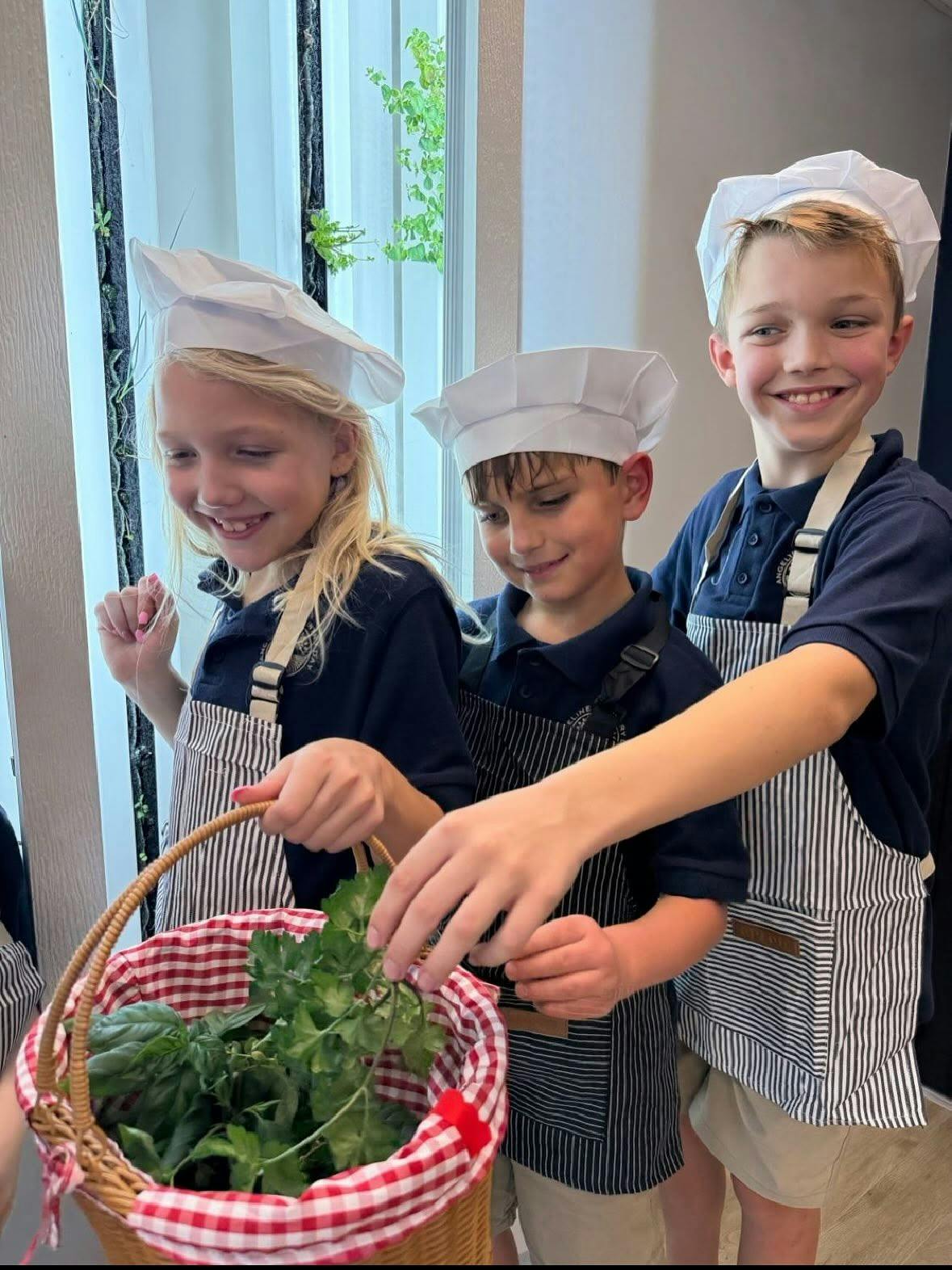 Three children in chef hats and aprons holding a basket of fresh herbs