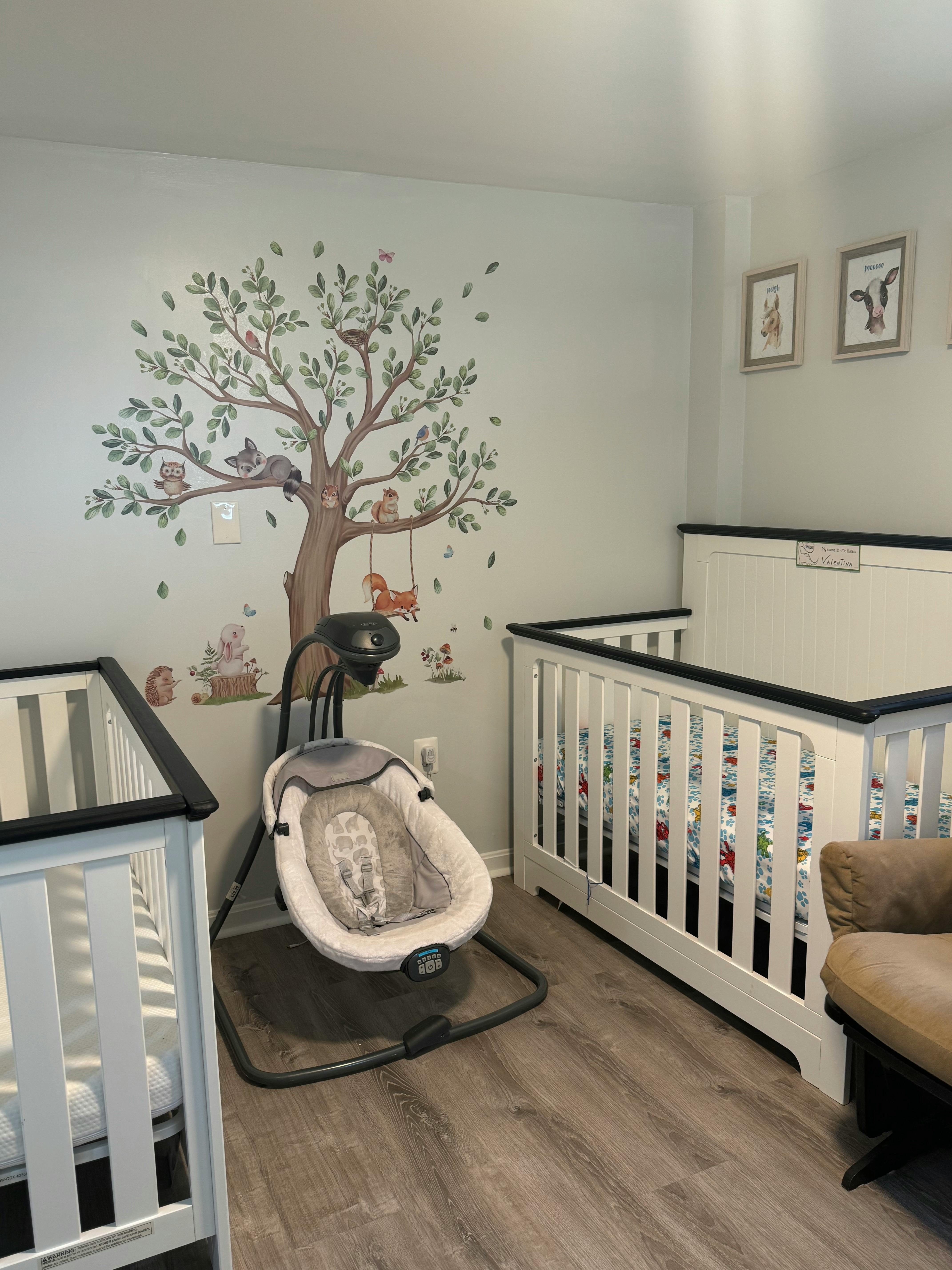 Nursery room with two cribs, a baby swing, and a tree wall decal