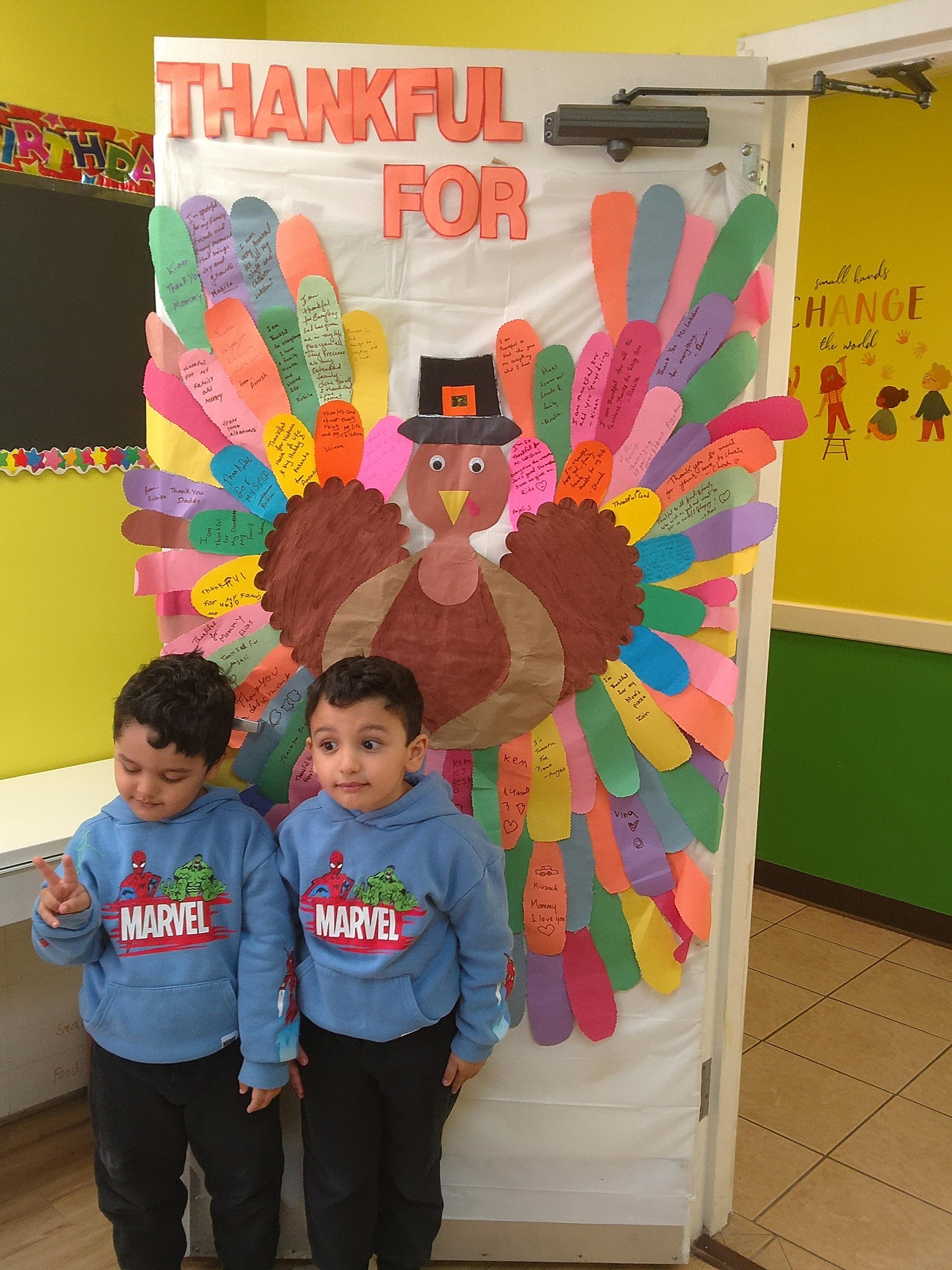 Two children standing in front of a colorful turkey decoration on a door with "THANKFUL FOR"