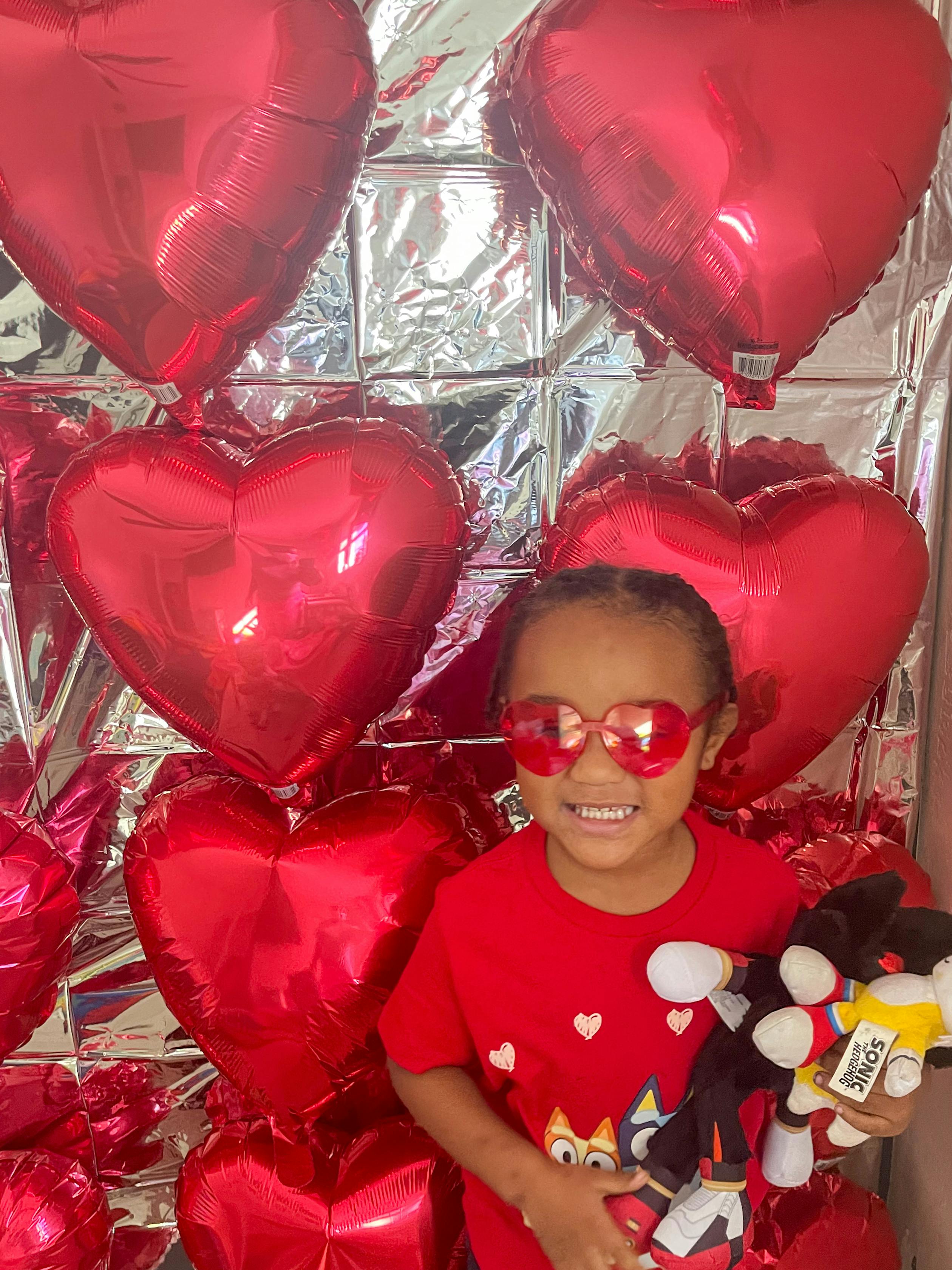 Child in heart-shaped glasses holding a plush toy, surrounded by red heart balloons