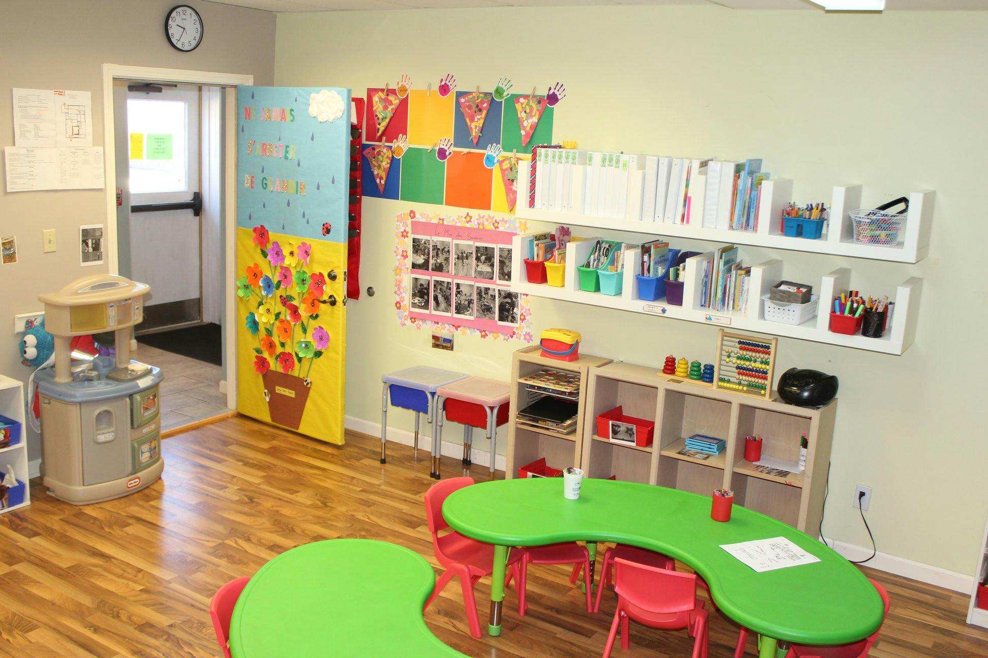 Classroom with green tables and red chairs, shelves with supplies, and colorful decorations