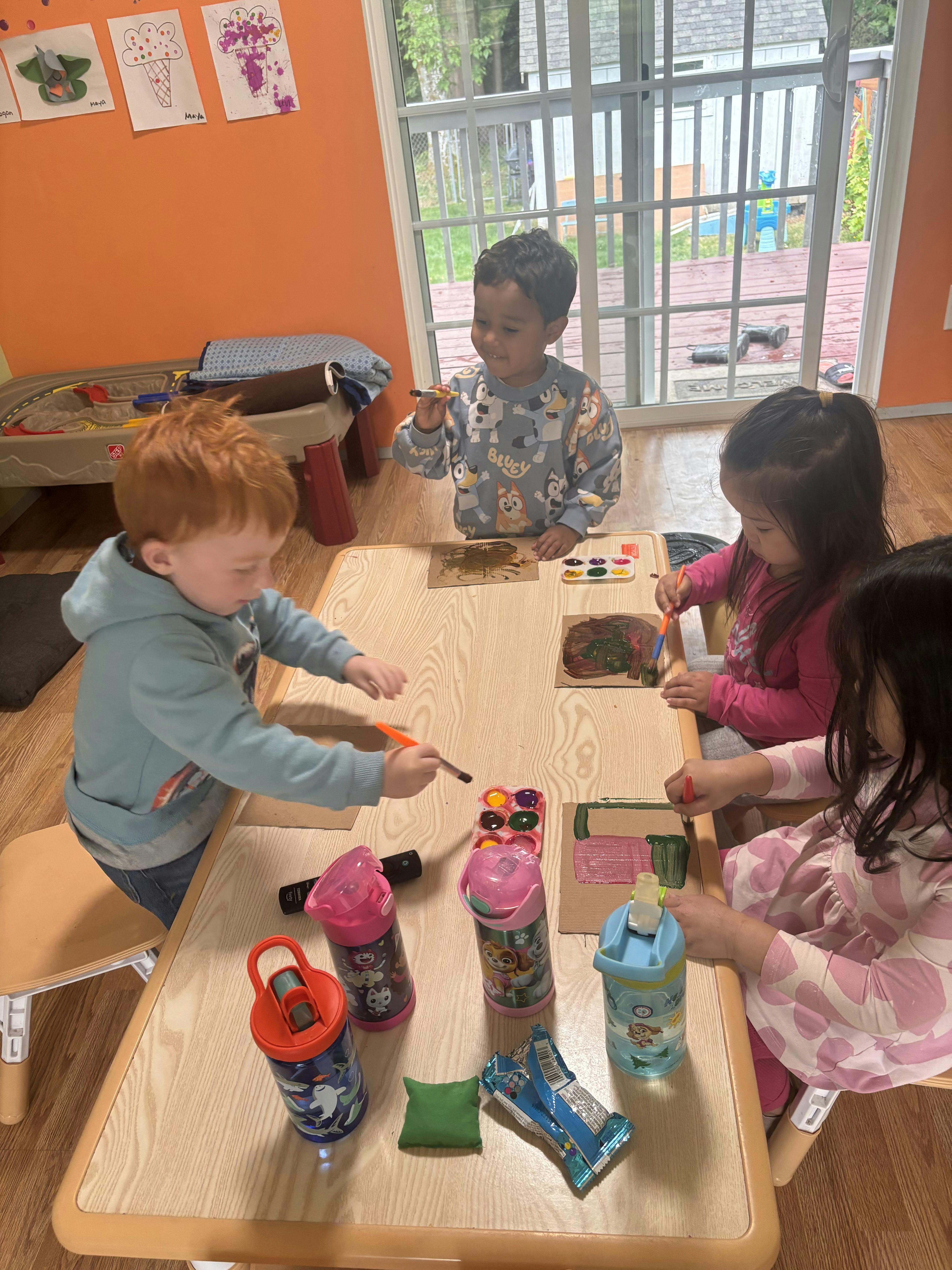 Four children painting on cardboard at a table in a classroom