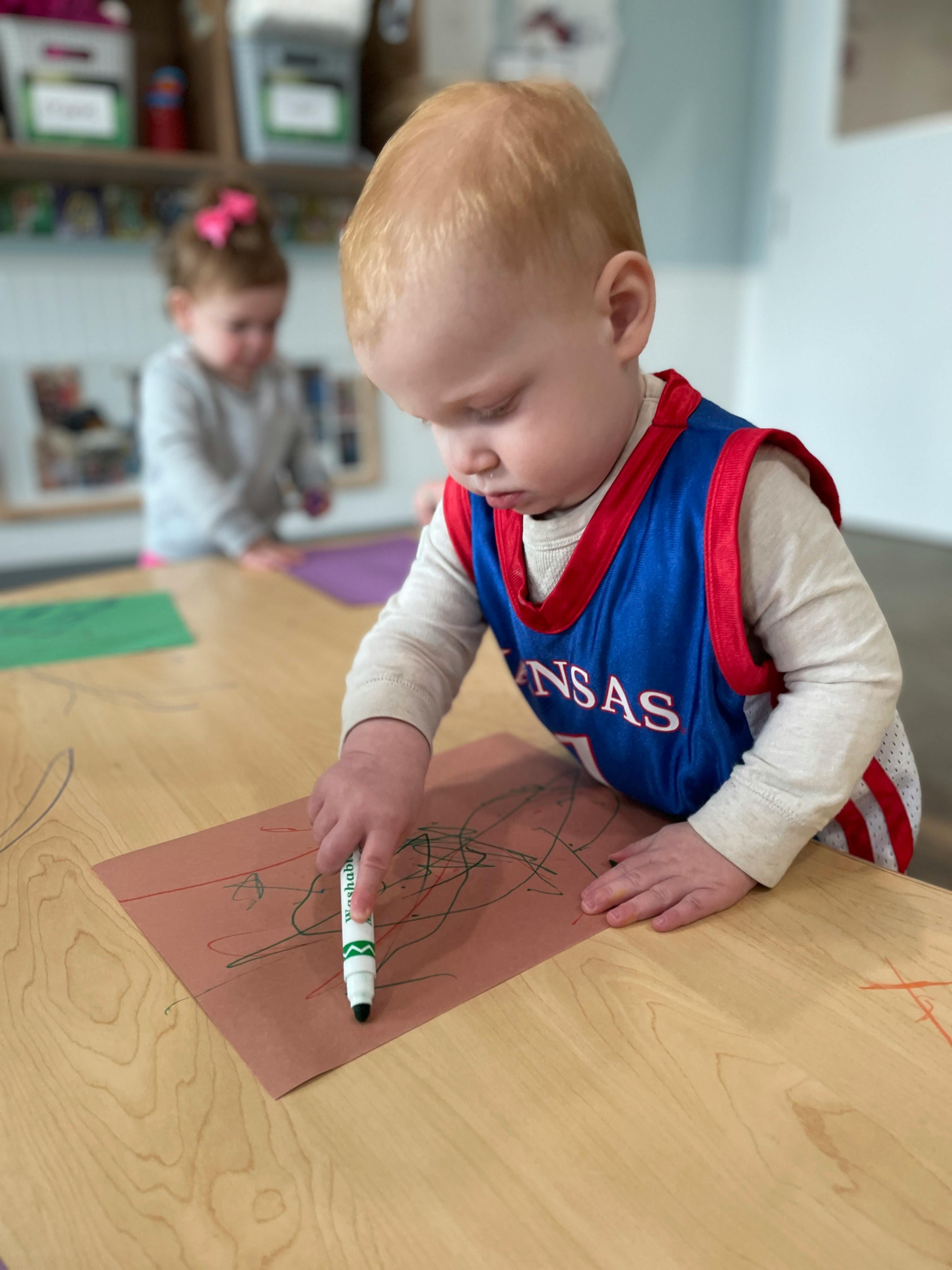 Child drawing on paper with a marker atable, another child in the background