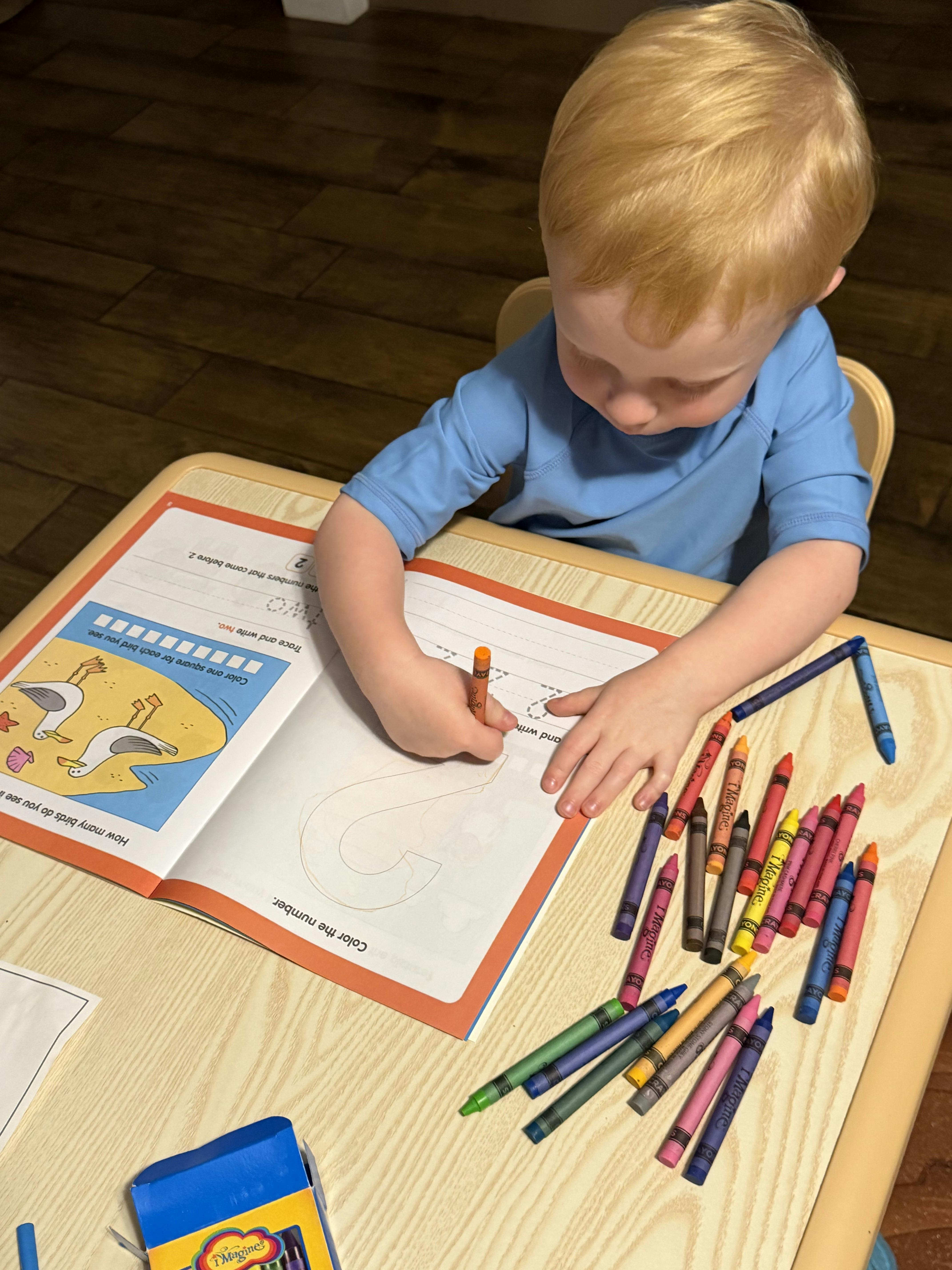Child coloring the number two in an activity book at a table with crayons