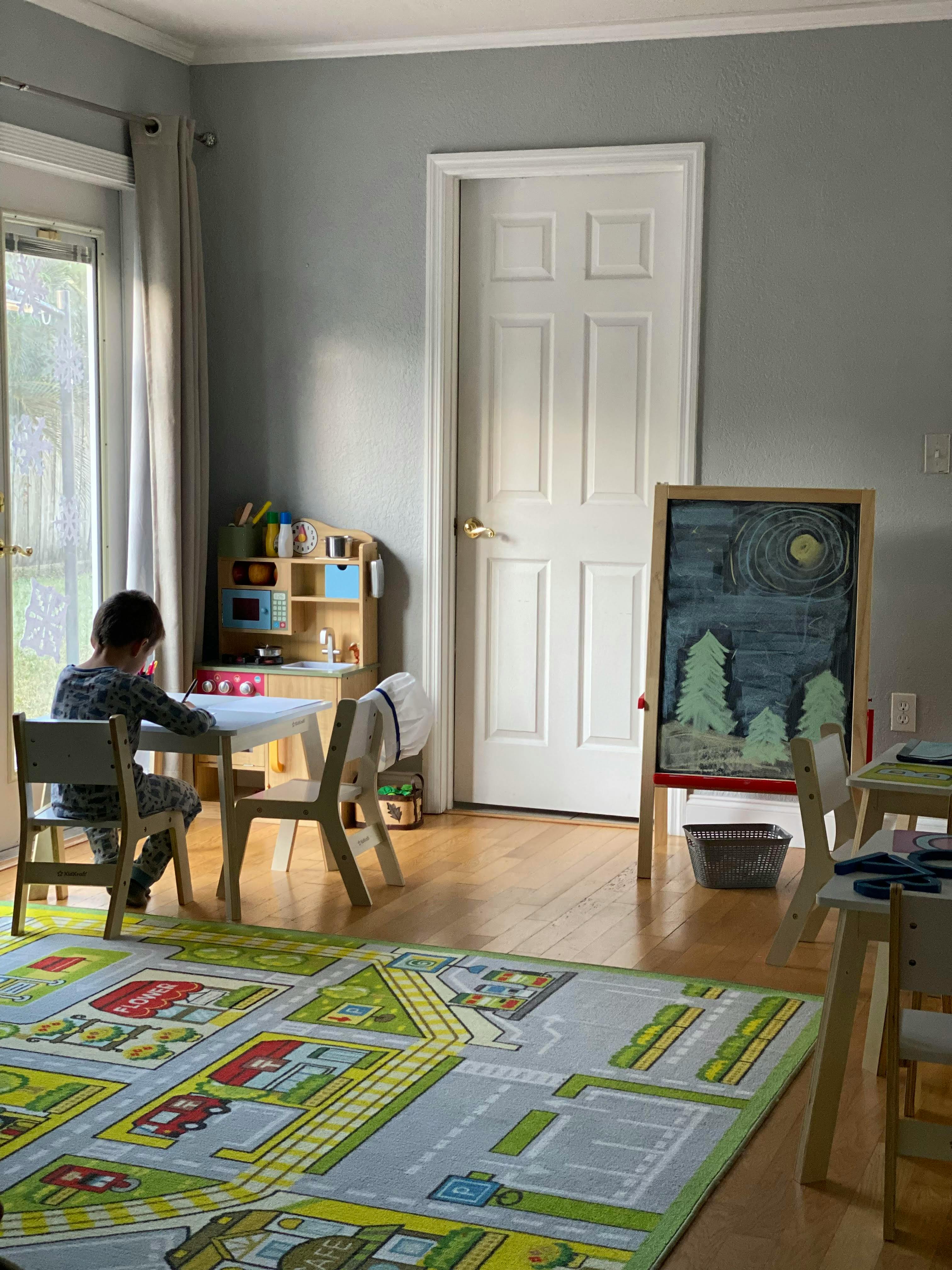 Child sitting at a small table in a room with a play kitchen and chalkboard