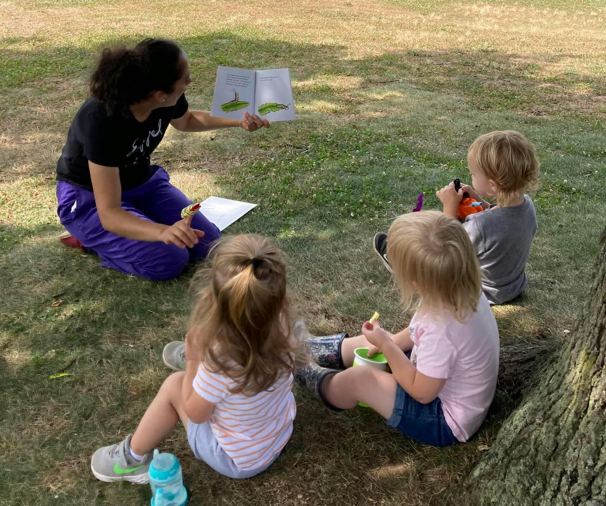 Person reading a book to three children outdoors