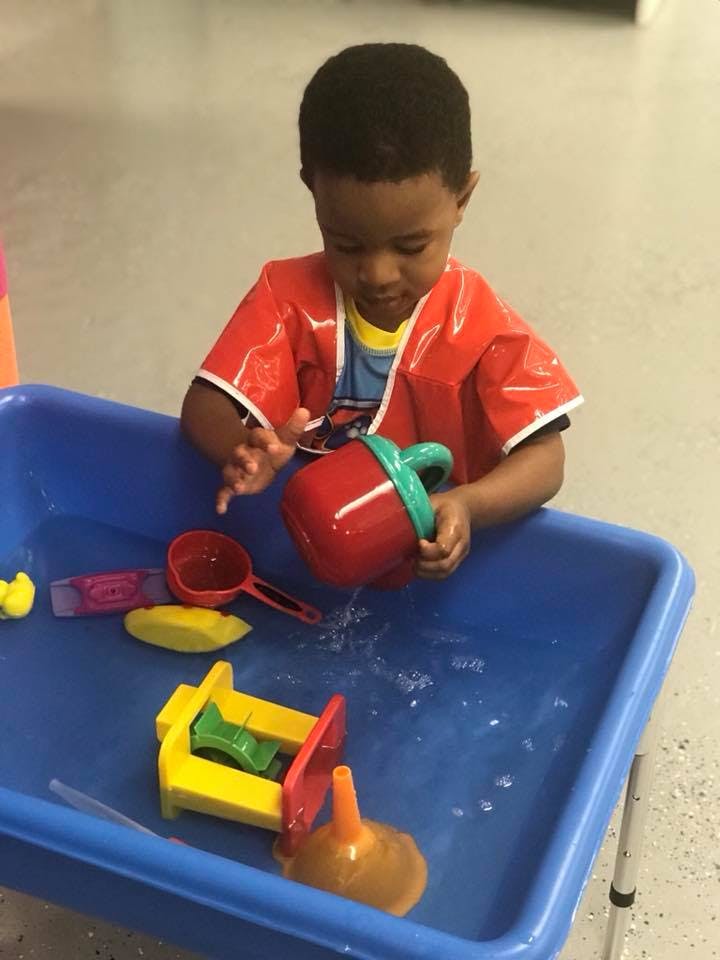 Child in a red smock playing with water and toys in a blue bin