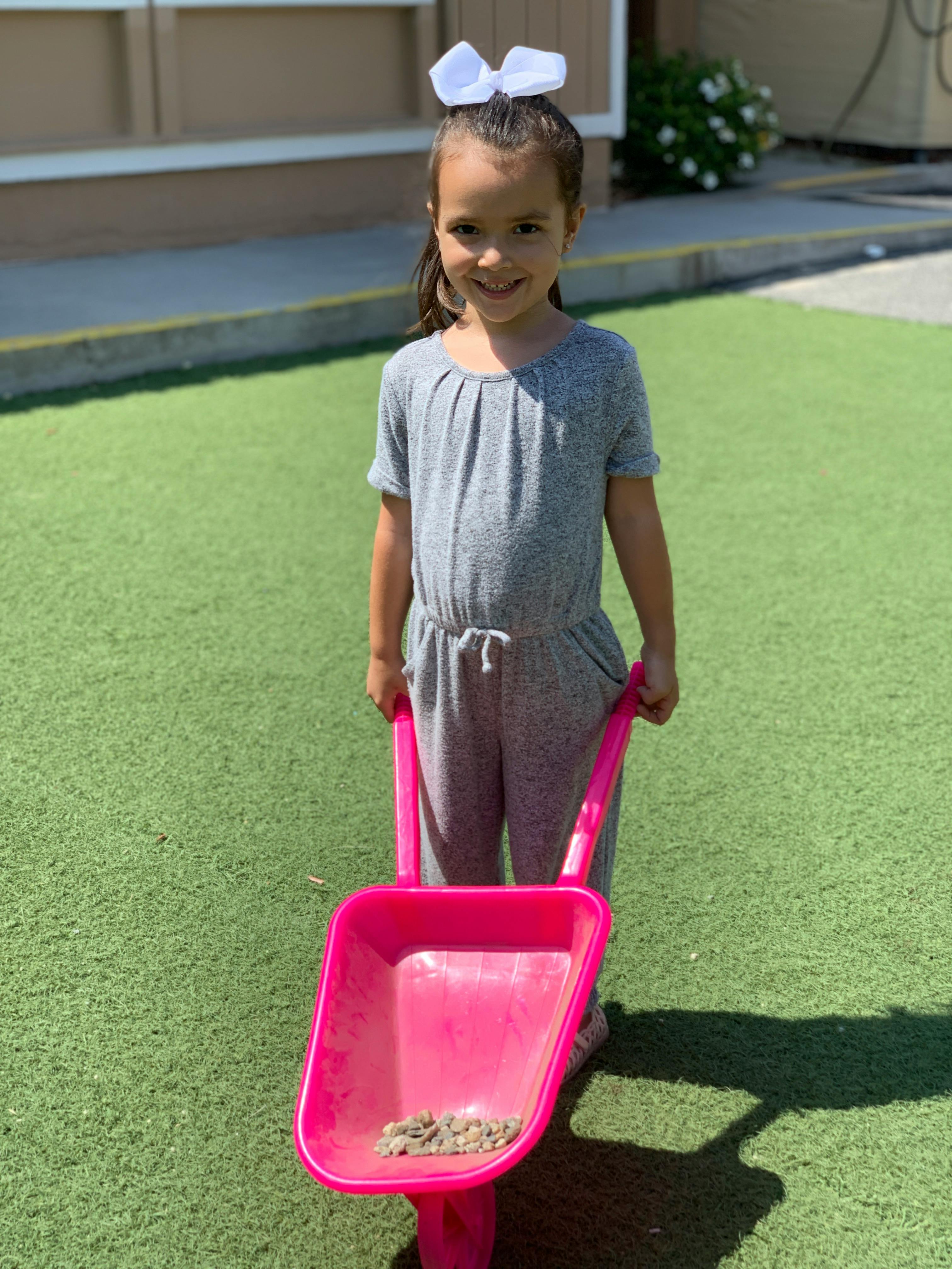 Child in gray jumpsuit holding a pink toy wheelbarrow
