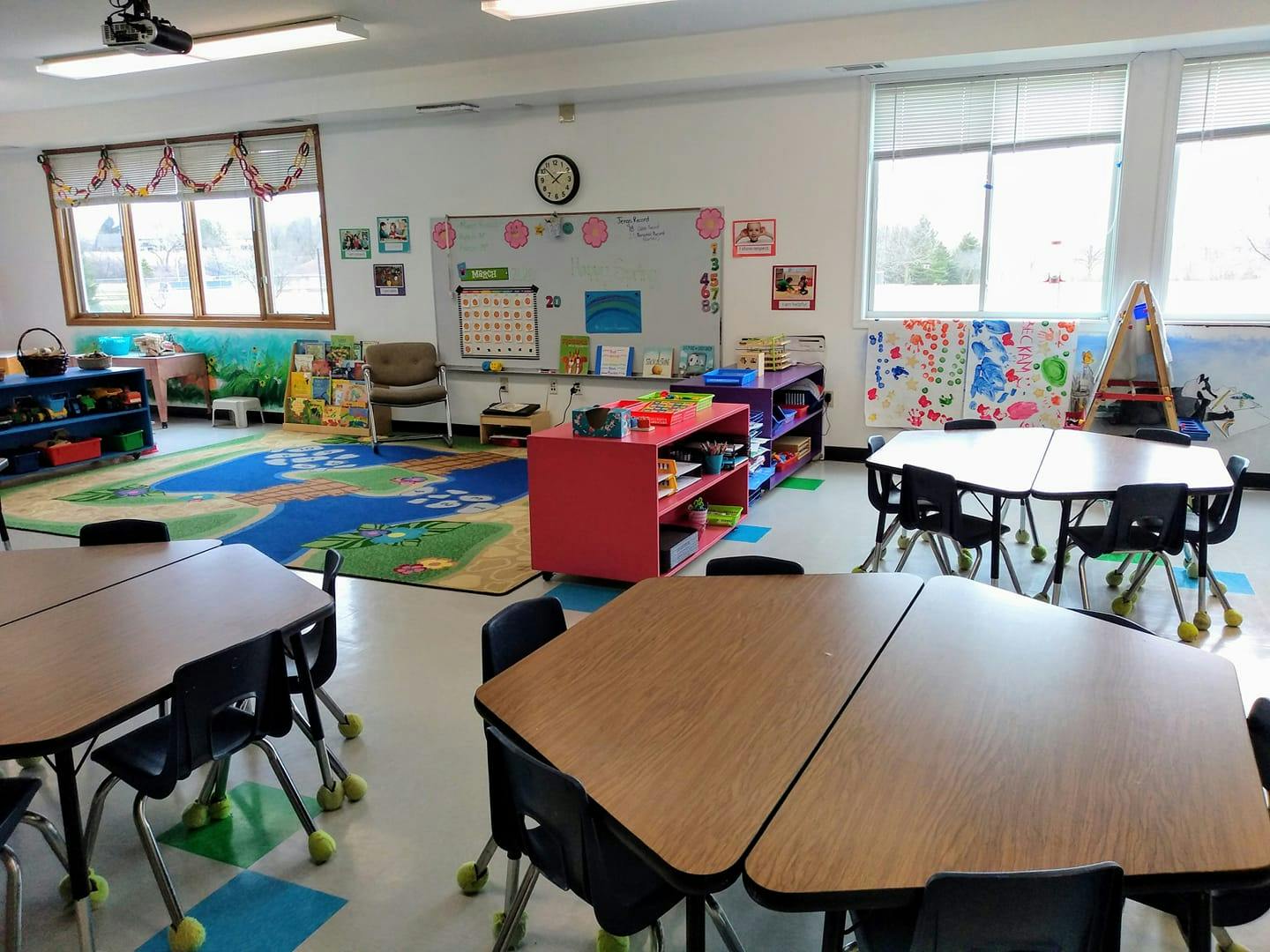 Empty classroom with small tables and chairs, a rug, and a whiteboard