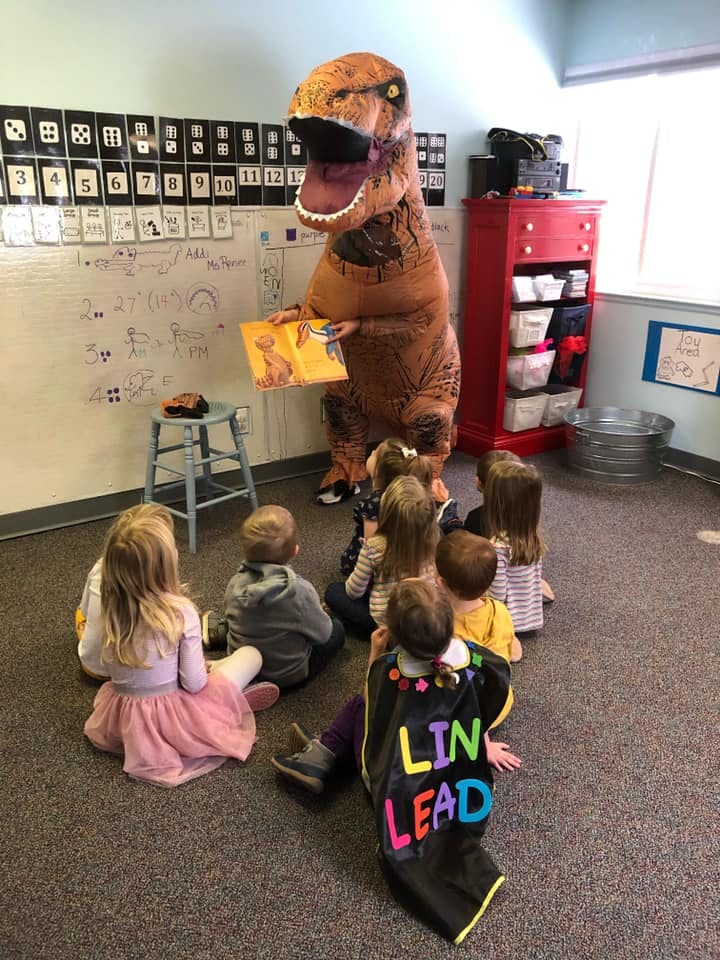 Person in dinosaur costume reading a book to children in a classroom