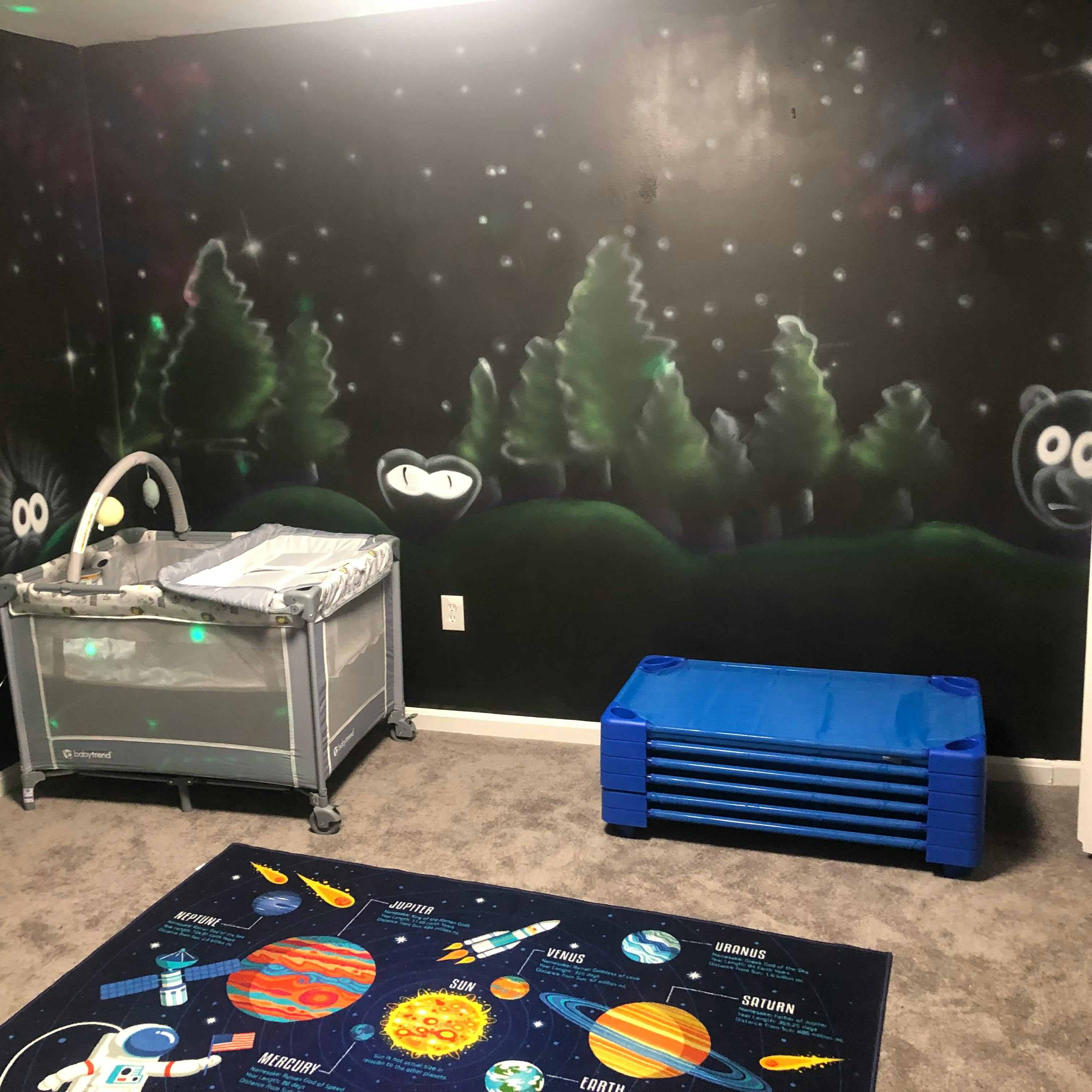 Playard, blue toddler bed, and space-themed rug in a room with a mural