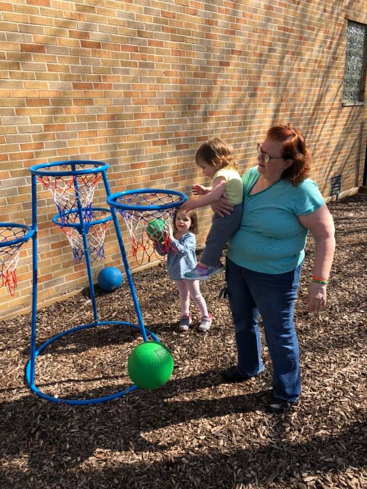 Person holding a child who is reaching for a basketball hoop, another child holds a ball