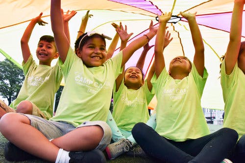 Children sitting with arms raised under a colorful parachute