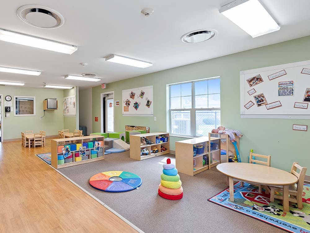 Classroom with small tables and chairs, shelves with toys, and colorful mat