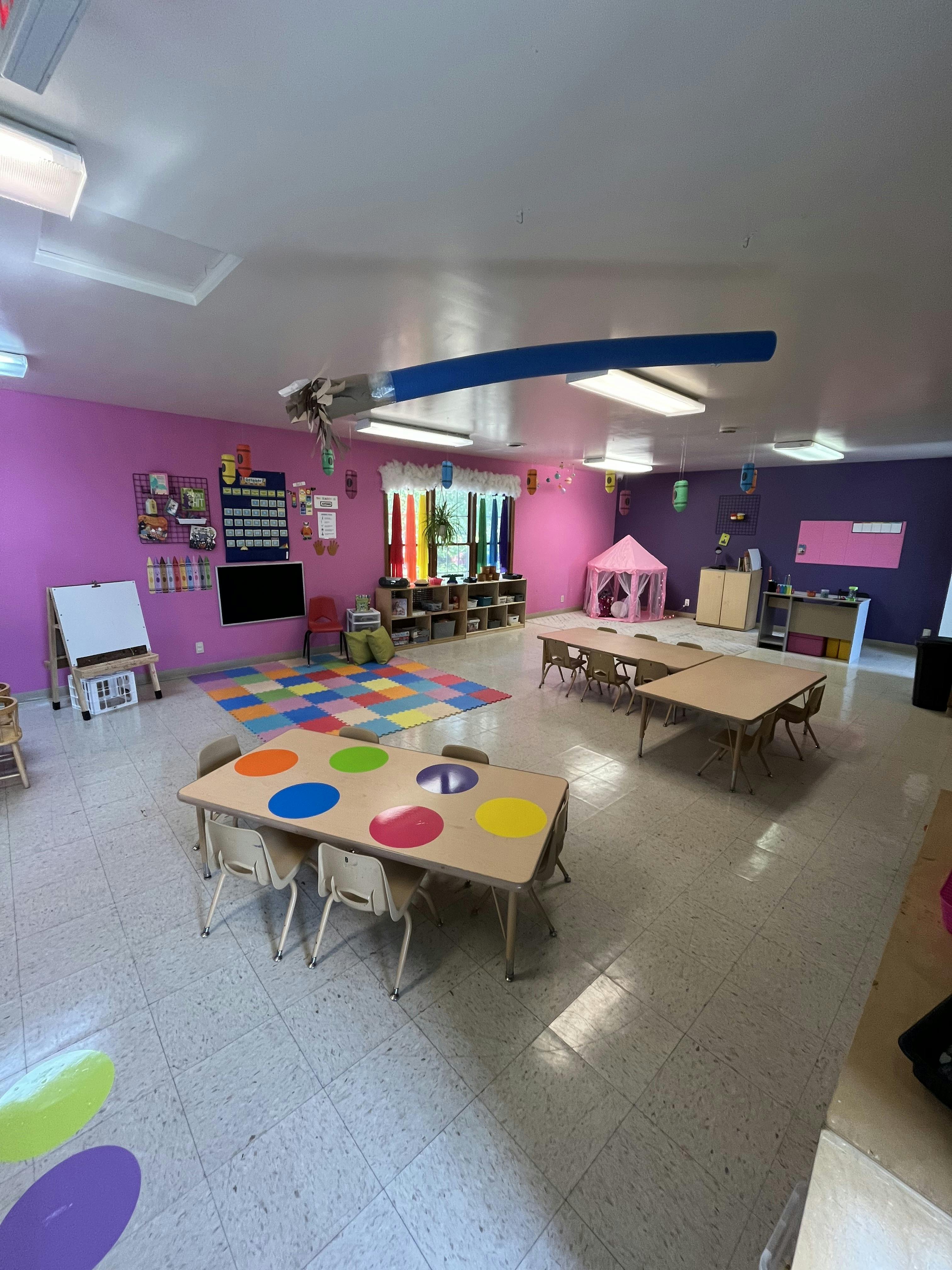 Classroom with small tables and chairs, colorful rug, and pink walls