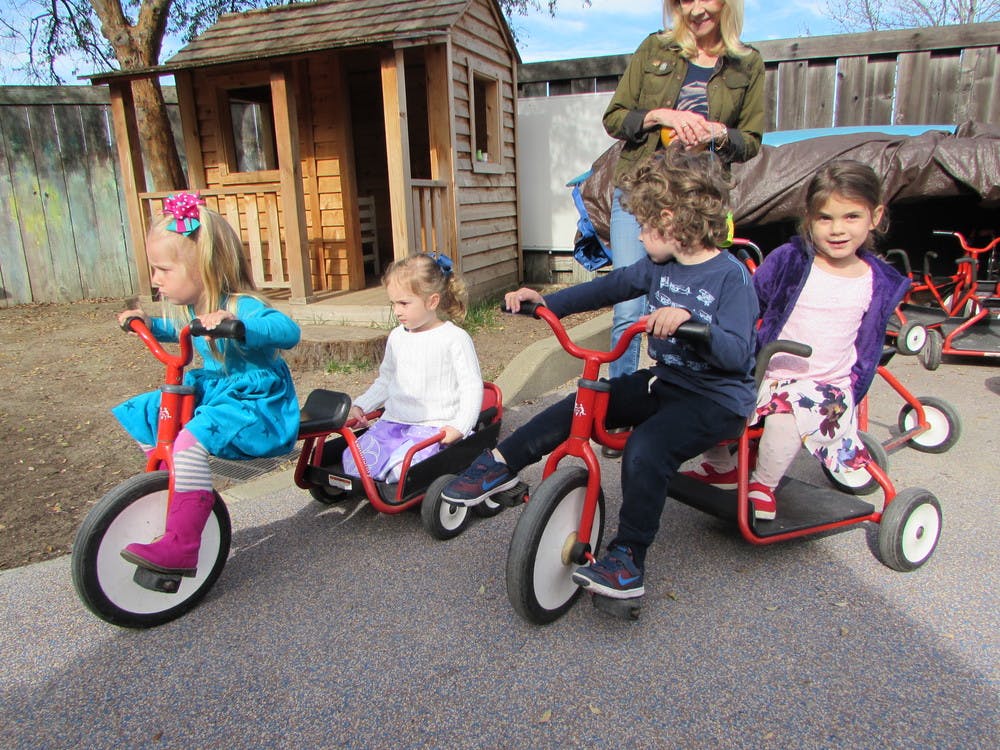 Three children riding tricycles outdoors with an adult standing behind them
