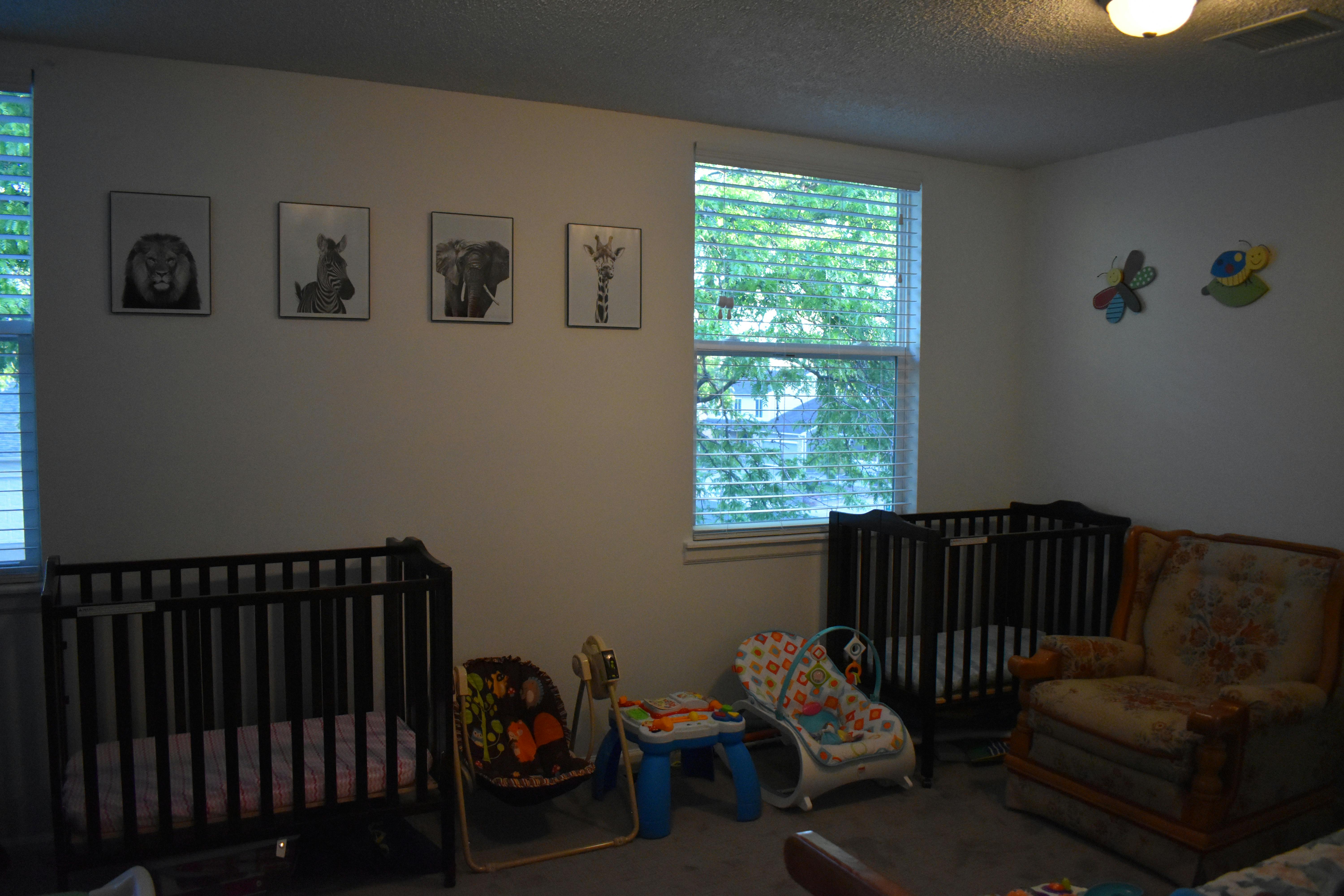 Two cribs in a room with windows and framed pictures on the wall