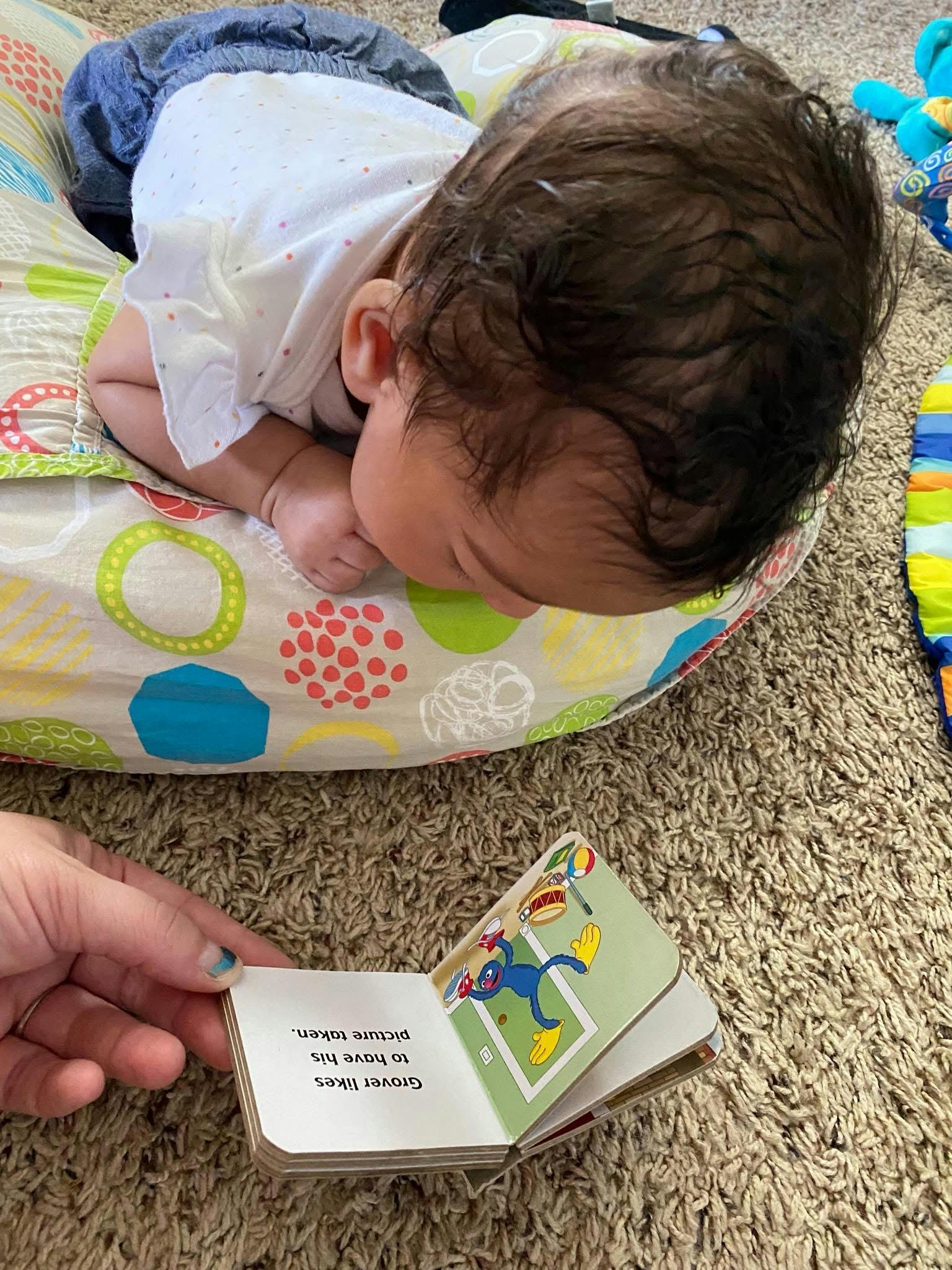 Baby lying on a pillow while a person reads a book with text "Grover likes to have his picture taken"