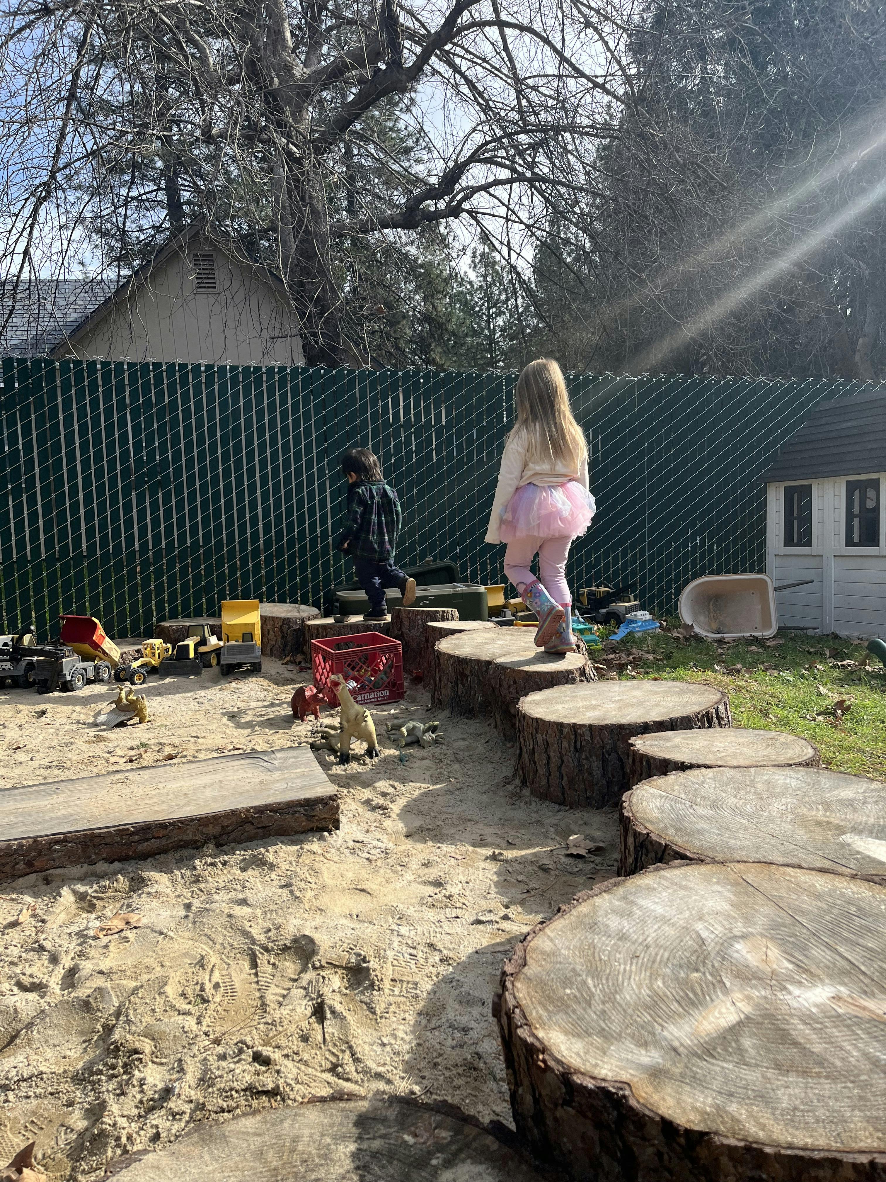Two children playing on tree stumps and in a sandbox with toy trucks