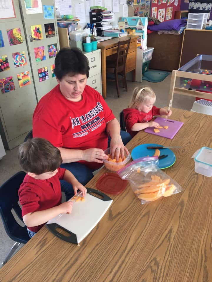 Adult and two children cutting food at a table in a classroom
