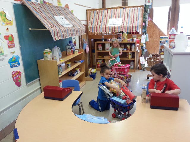 Three children playing in a classroom with toy shopping carts and shelves