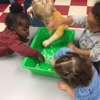 Four children playing with small objects in a green bin