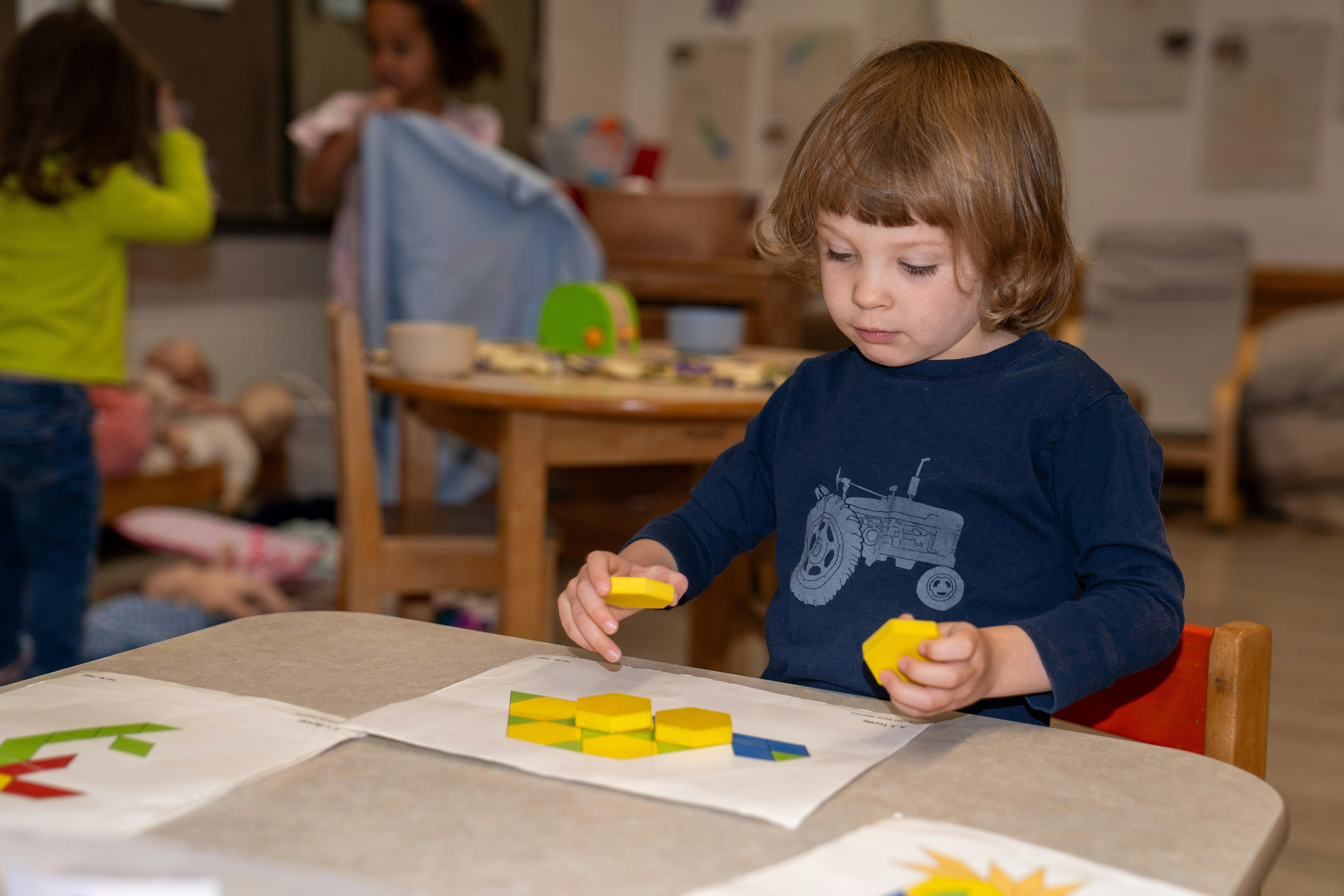 Child playing with yellow and blue shapes at a table in a classroom