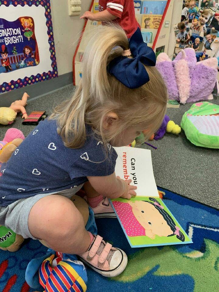 Child with blonde ponytail and blue bow reading a book on the floor
