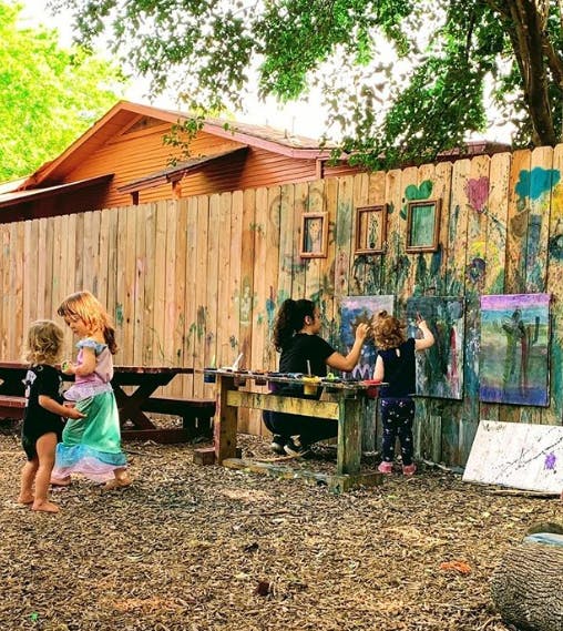 Adult and child painting at easels outdoors, two children standing nearby