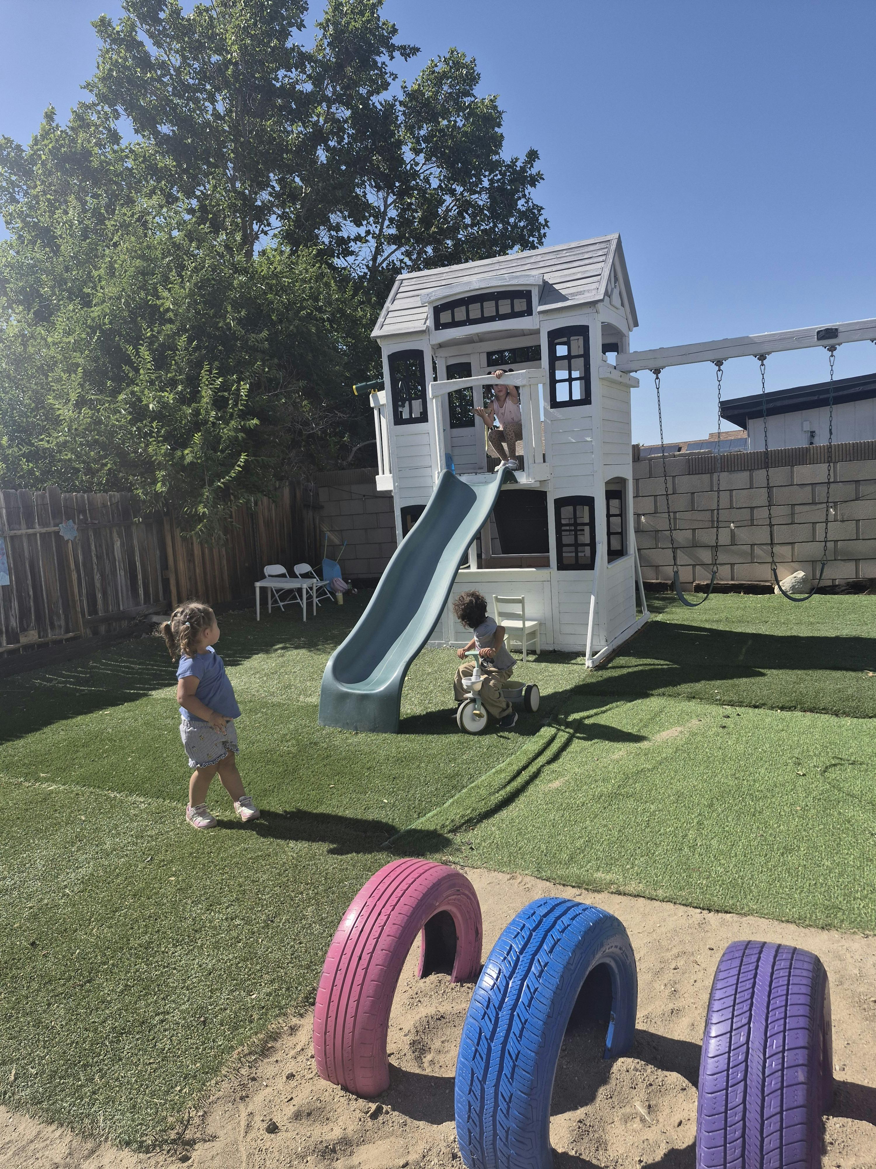 Child playing in a playhouse, another on a tricycle, and a child standing nearby
