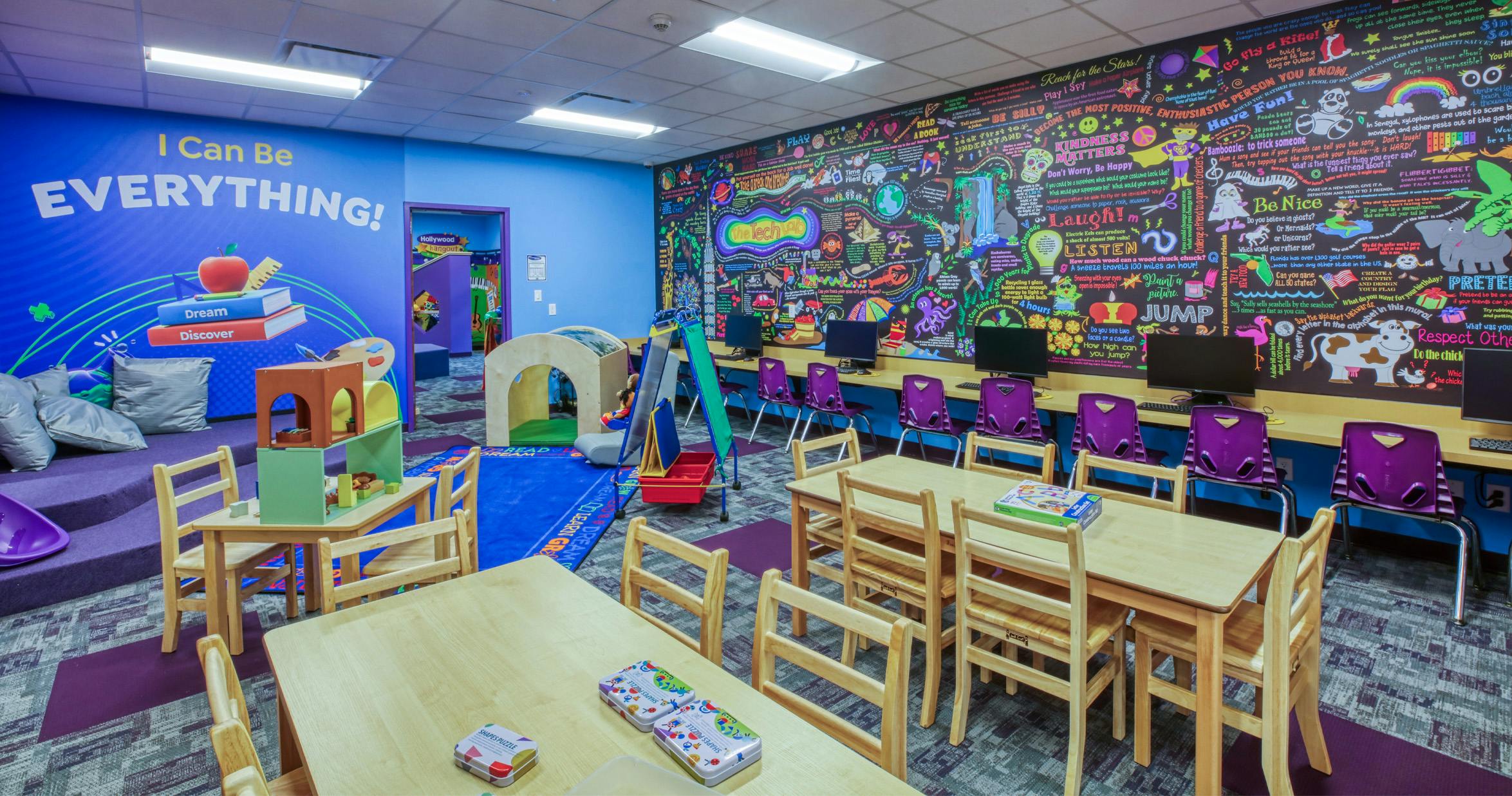 Colorful classroom with tables, chairs, computers, and a wall mural with text