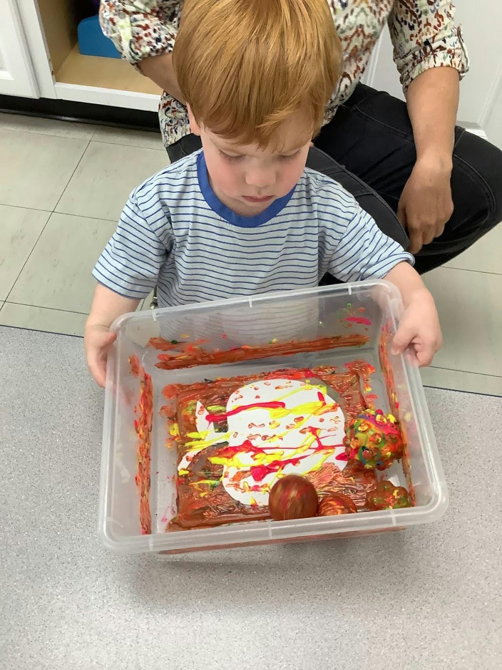 Child holding a plastic bin with painted balls and paper inside
