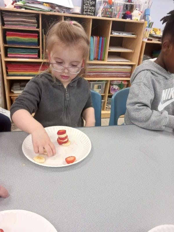 Child in glasses making a snack of fruit slices in a classroom