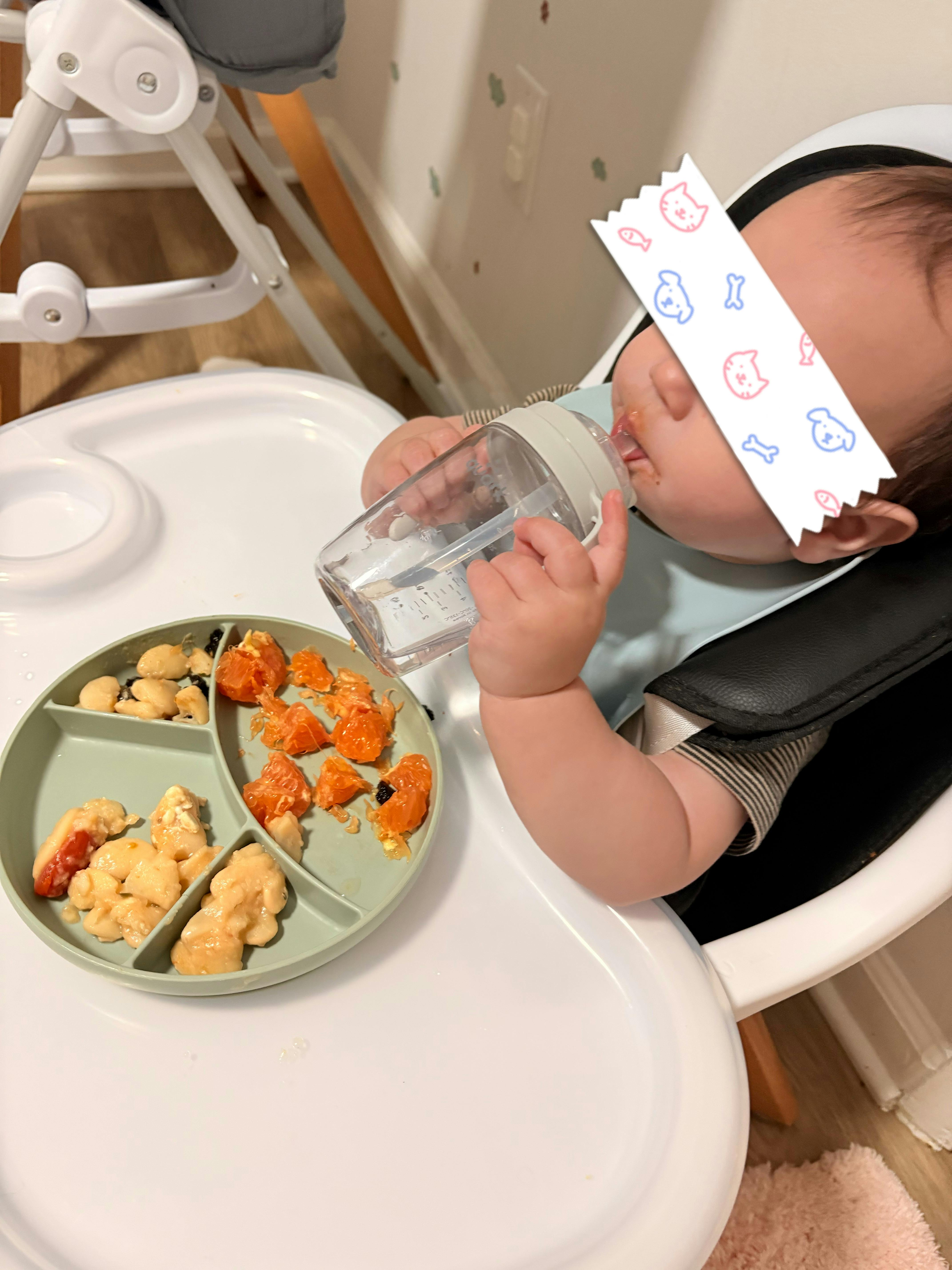 Child sitting in a high chair drinking from a water bottle near a food tray