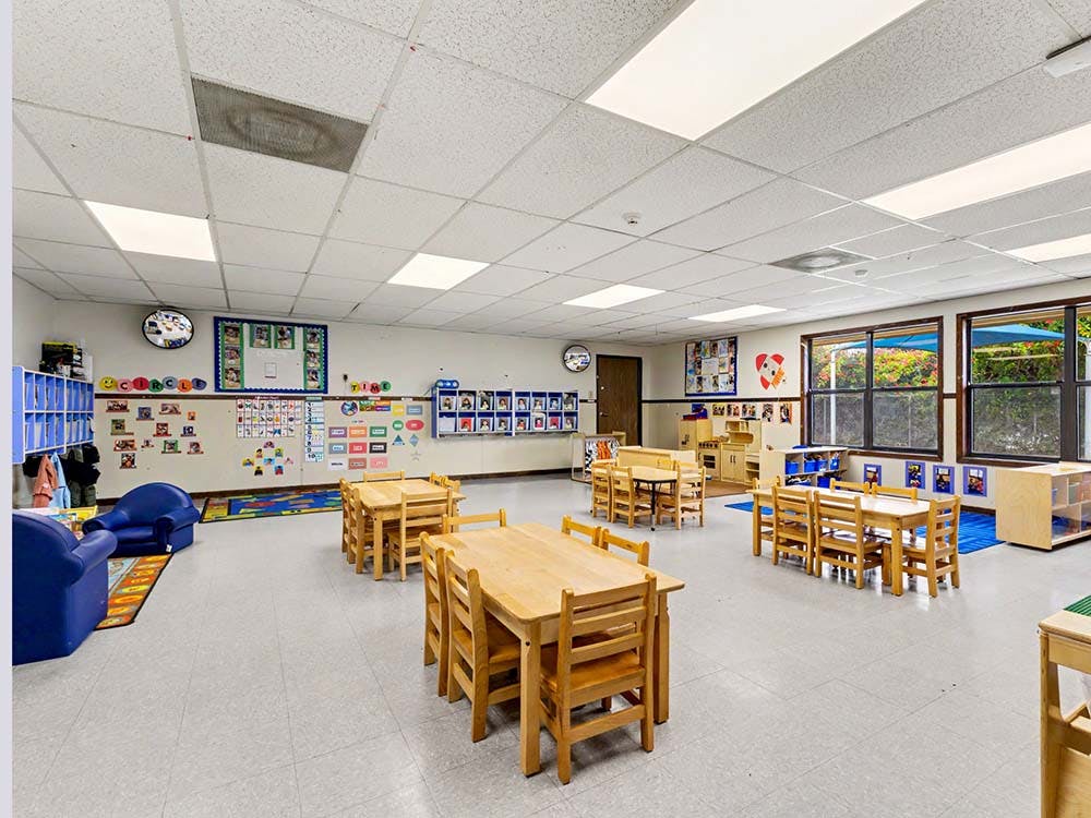 Empty preschool classroom with small wooden tables and chairs, and blue armchairs