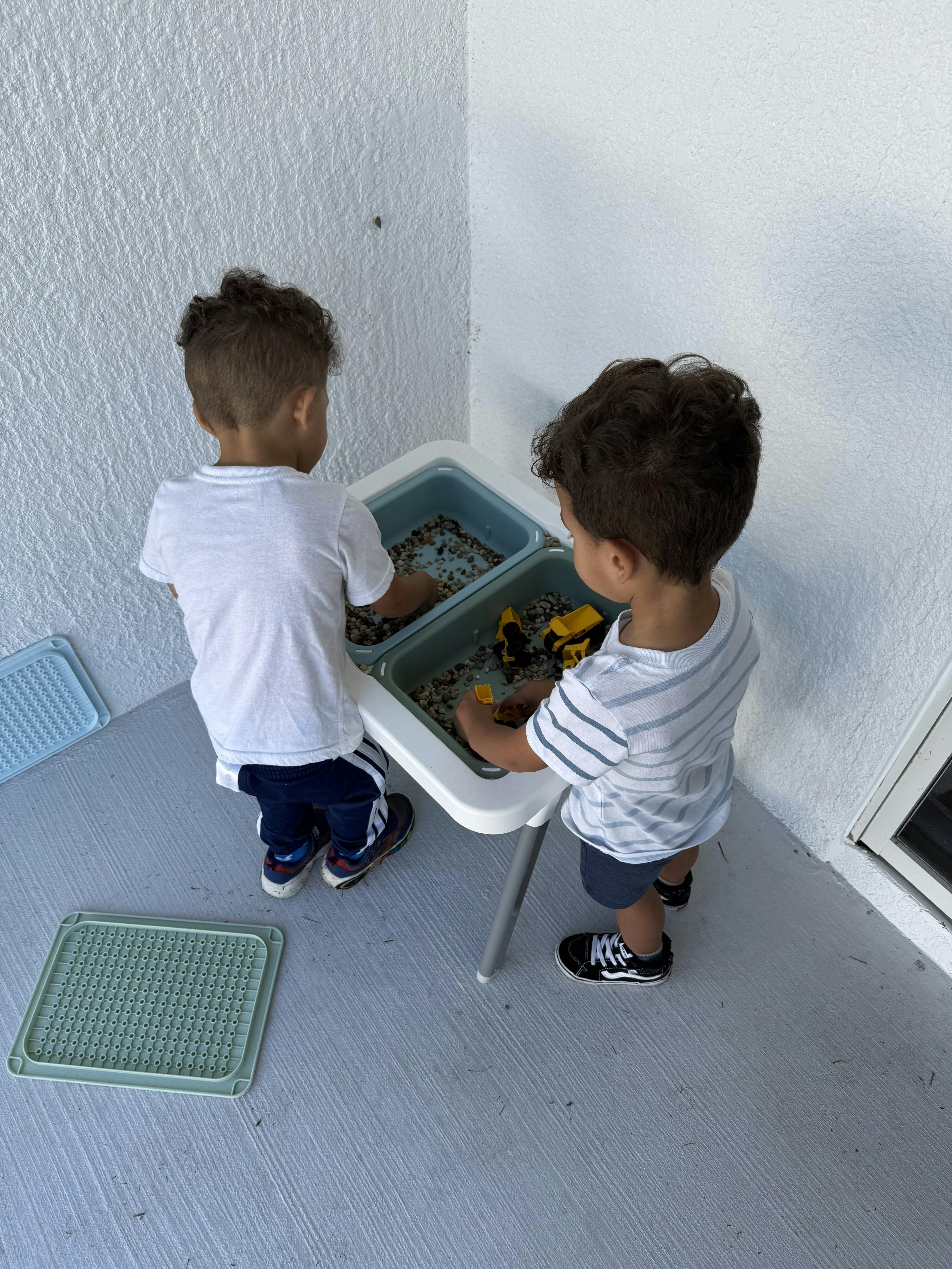 Two children playing at a water table with pebbles and toy construction trucks
