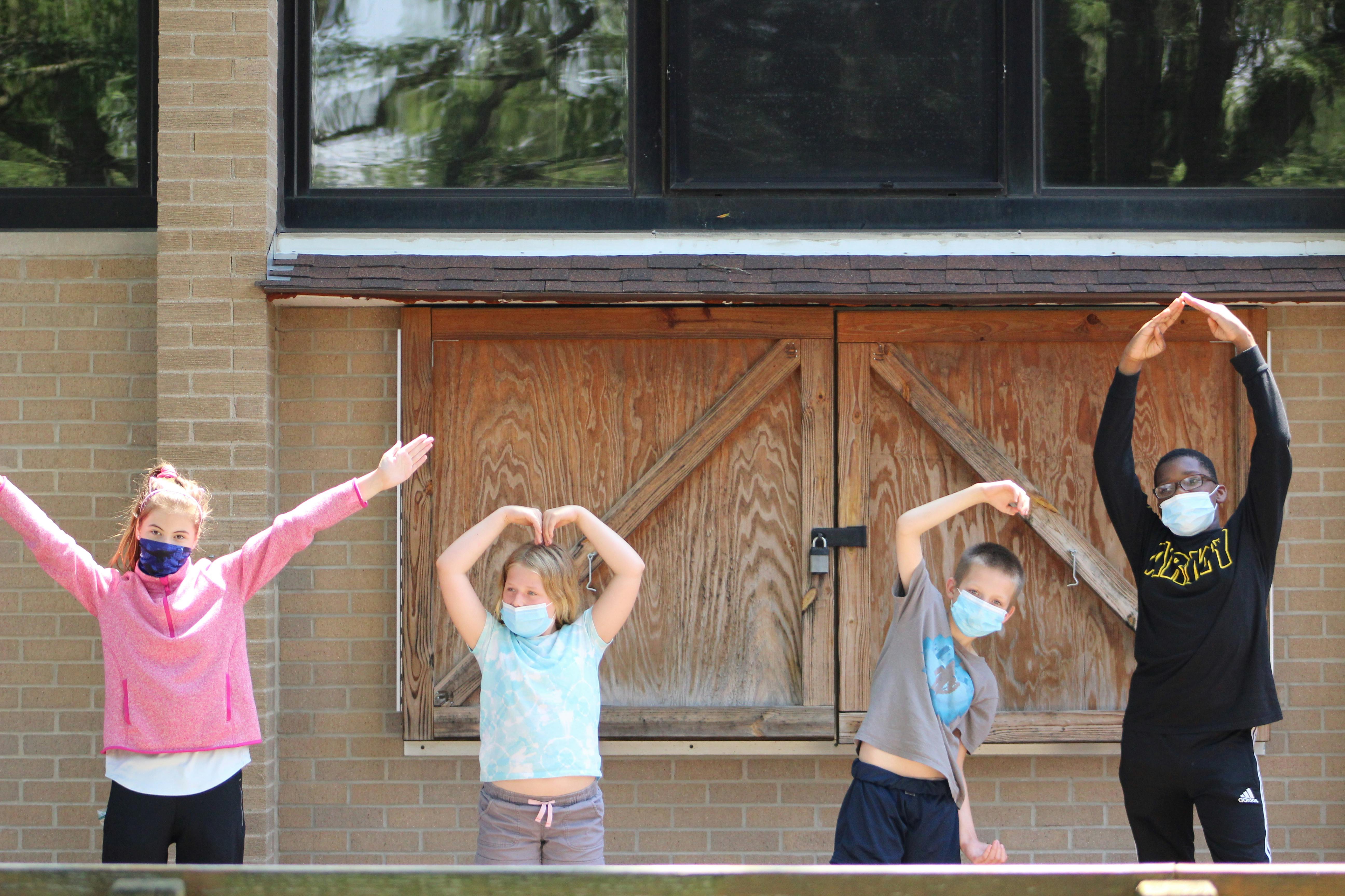 Four children wearing masks making letter shapes with their arms